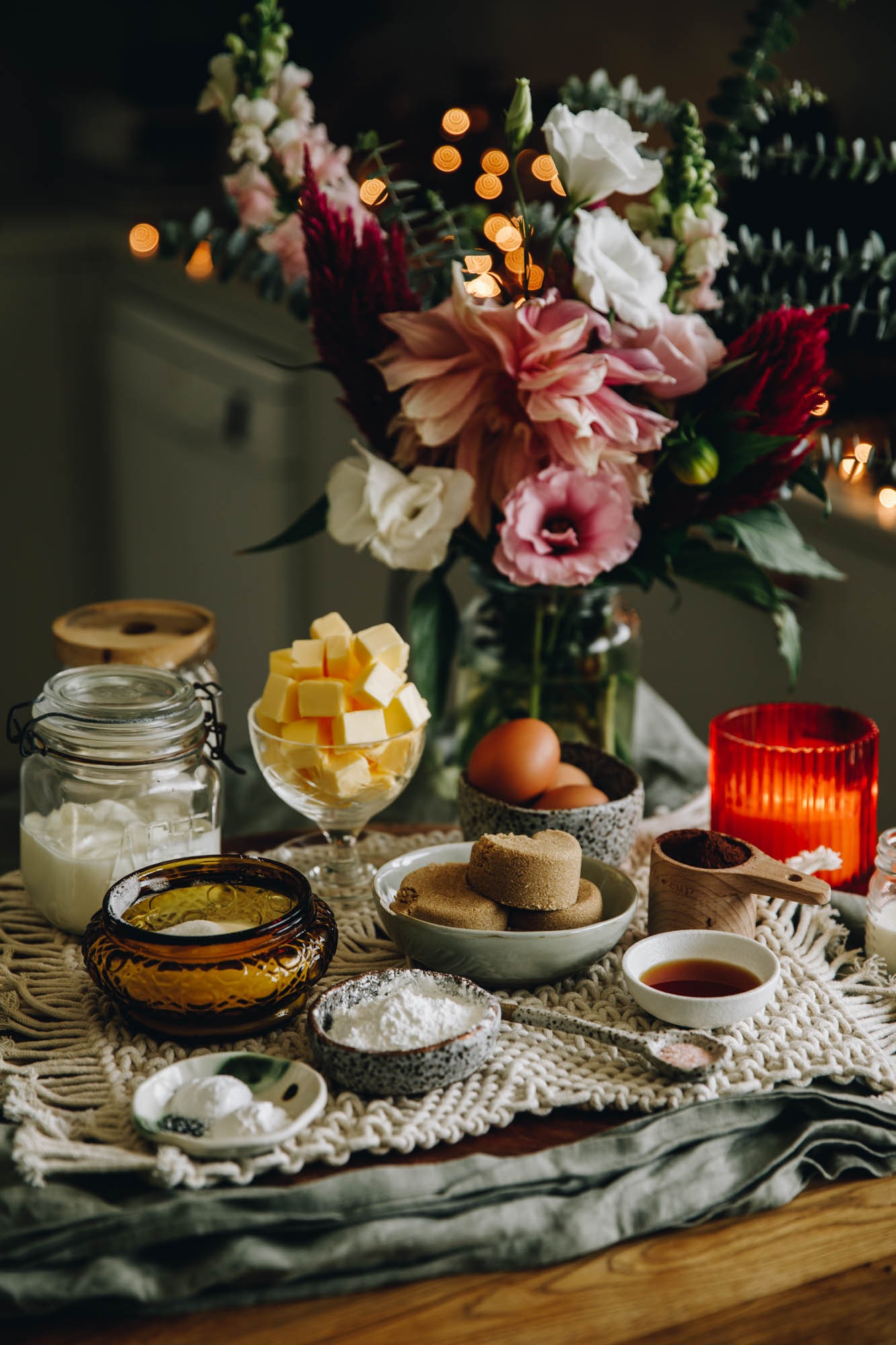 A weaved cream mat sits on a blue table cloth, on a wooden table. On the mat is ingredients in vintage bowls and ramekins. Cubed butter, brown sugar, eggs, flour, vanilla, salt, a glass jar, an orange burning candle and a vase of large flowers are in view. Golden fairy lights are in the background.