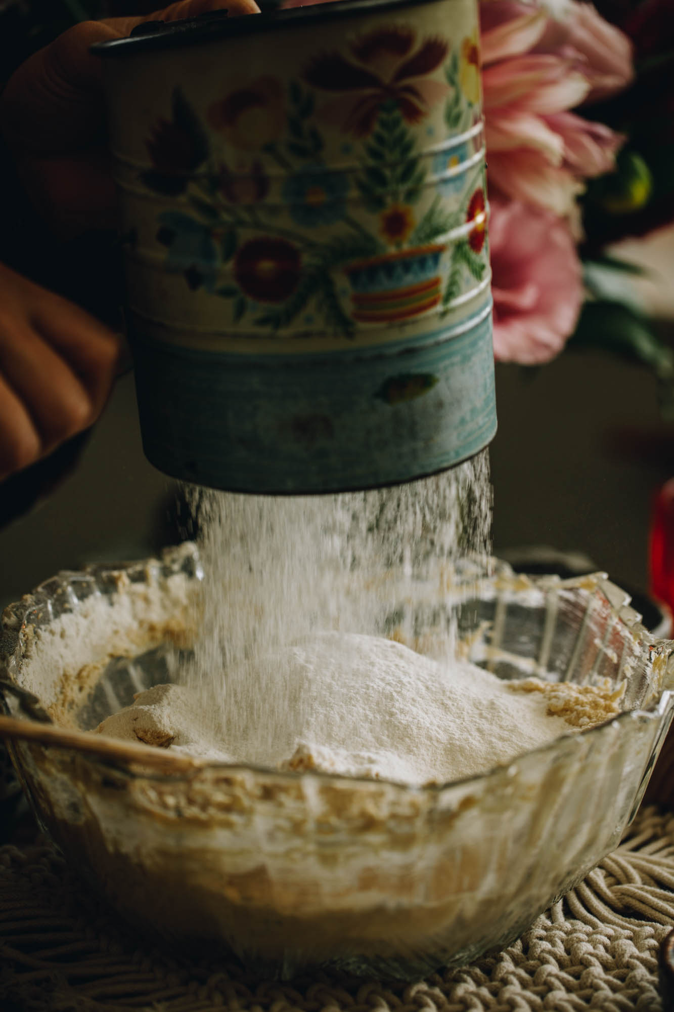 A glass vintage bowl sits on a weaved cream mat. In the bowl is whipped butter and Naomi using a blue vintage sifter to sift the dry ingredients on top it. Pink flowers are in the background.