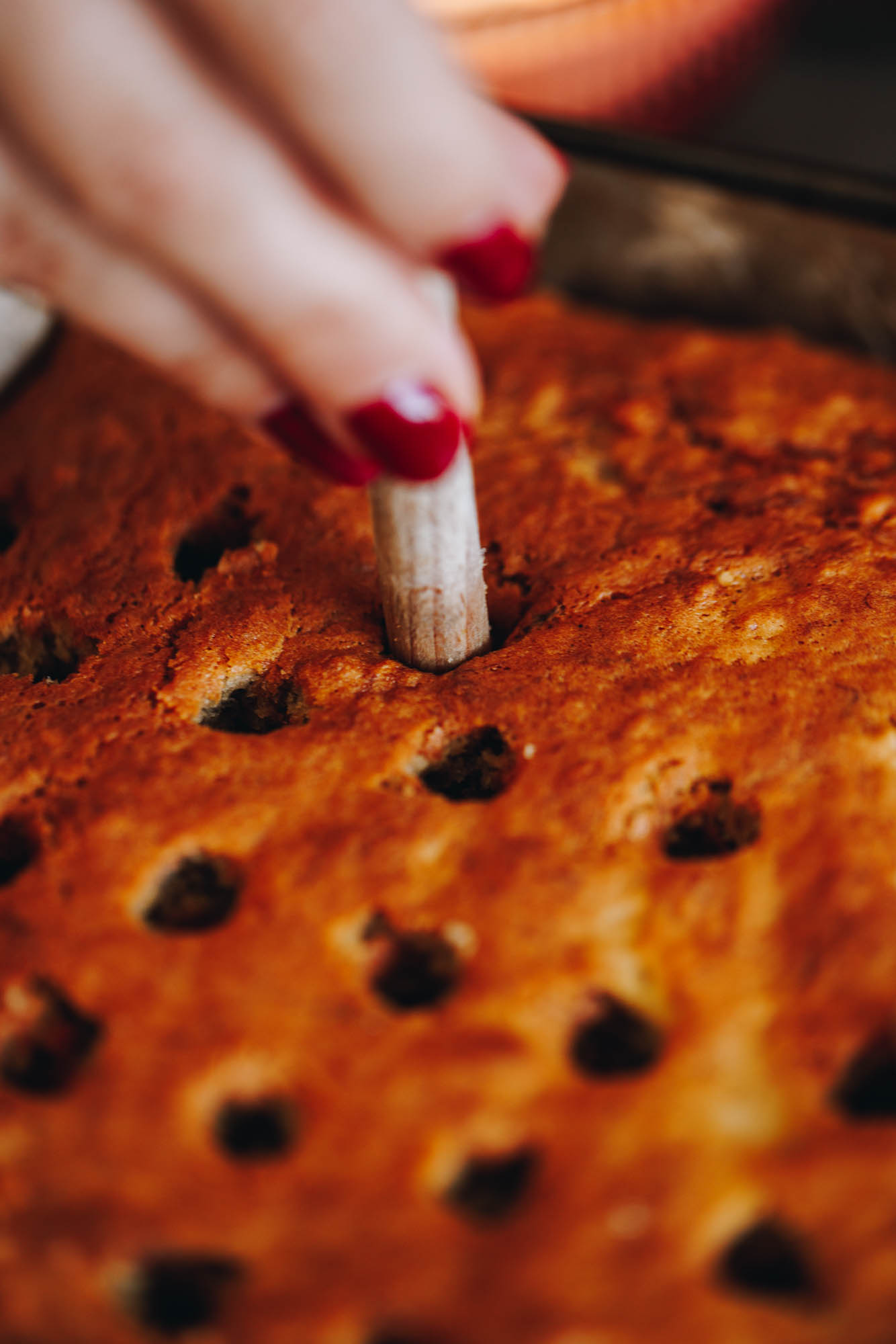 A golden baked cake has holes all over it and a wooden handle is poking a hole in to the centre of the cake. 