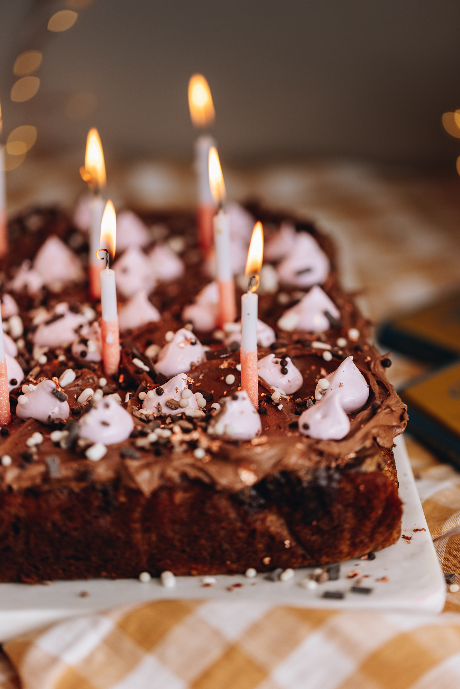 A freshly baked Chocolate ganache poke cake is on a white board. The cake has been decorated with a chocolate buttercream and blobs of pink meringue. Brown and white sprinkles are also on it. White and pink candles are lit on the cake too. 