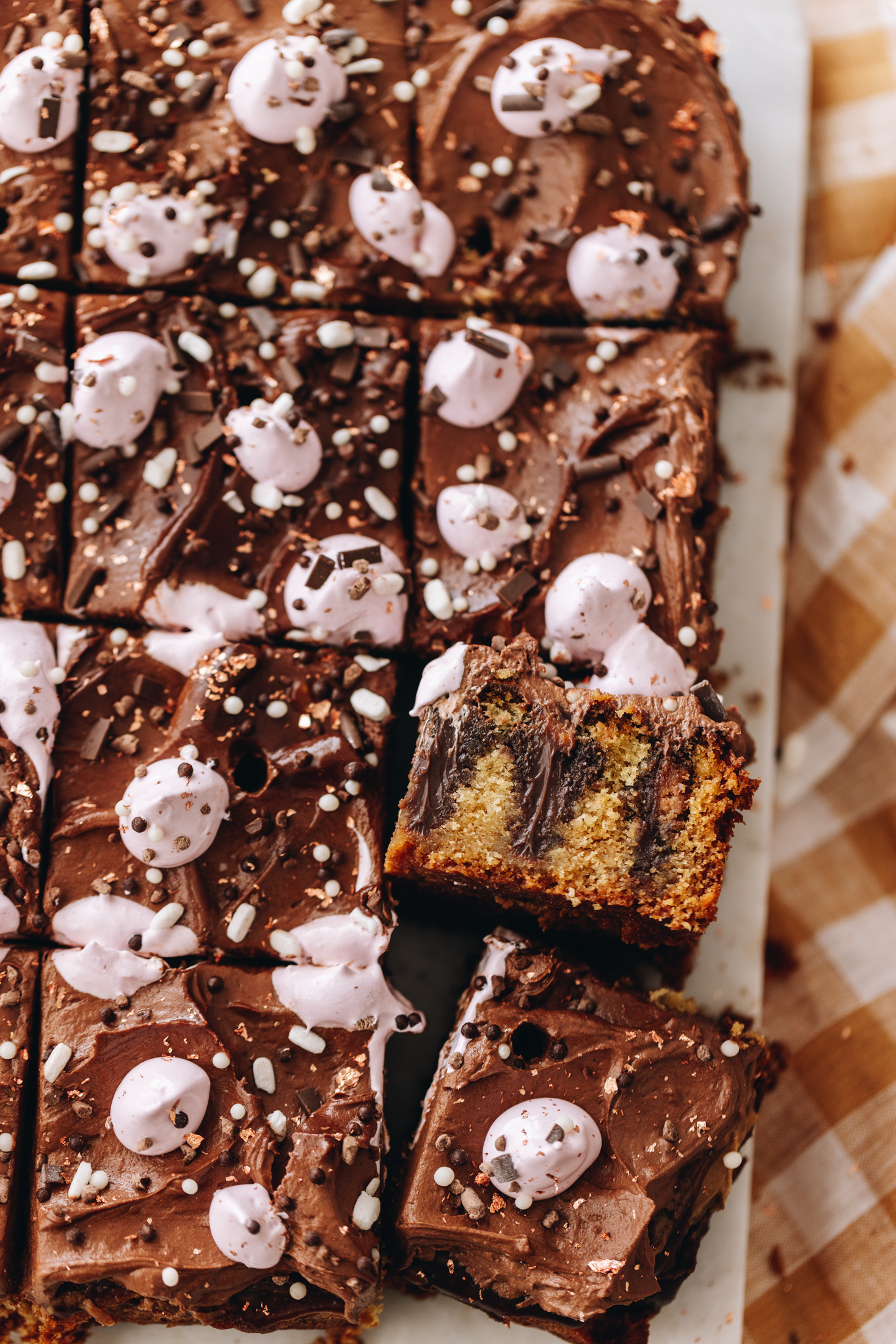 A flat lay shot shows a fresh decorated chocolate ganache poke cake. The cake has been in to squares and ons piece is flipped over. The slice shows the vanilla cake with chocolate ganache poked in to it. It is topped with a chocolate buttercream and pink blobs of meringue.
