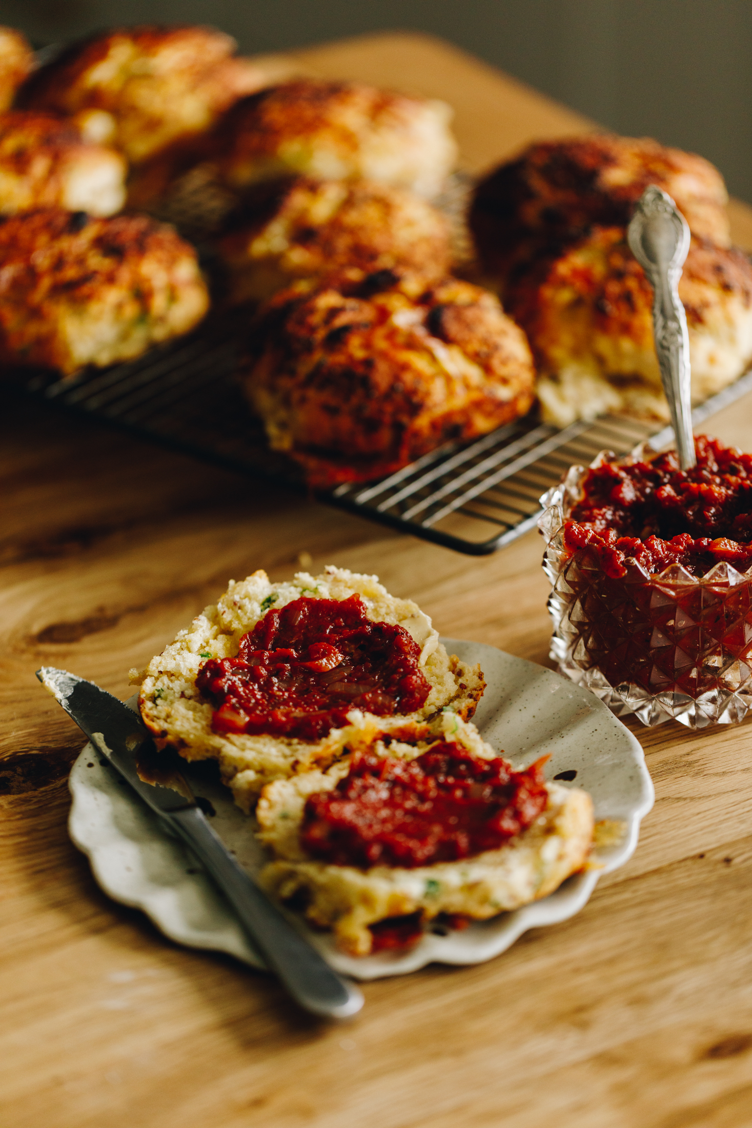 On a wooden table sits a ceramic crinkled edge plate with cheese scones on it opened up and topped with an easy tomato chutney recipe. Behind the scones is a jar of the chutney and cheese scones on a wire rack are behind the jar. 