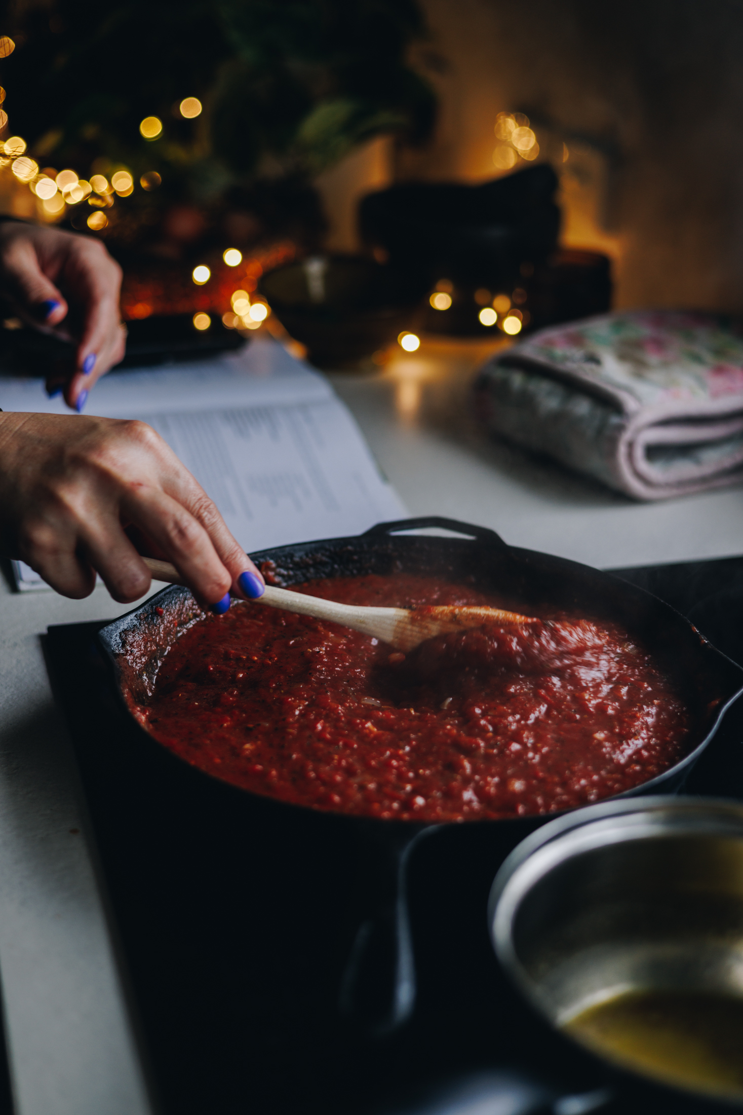 A black cast iron pan is on a black stove with an easy tomato chutney recipe cooking in it. Naomi is stirring with a wooden spoon. Next to the pan is a cookbook open and fairy lights in the background. On the other side of the pan is a pot with melted butter in it. 