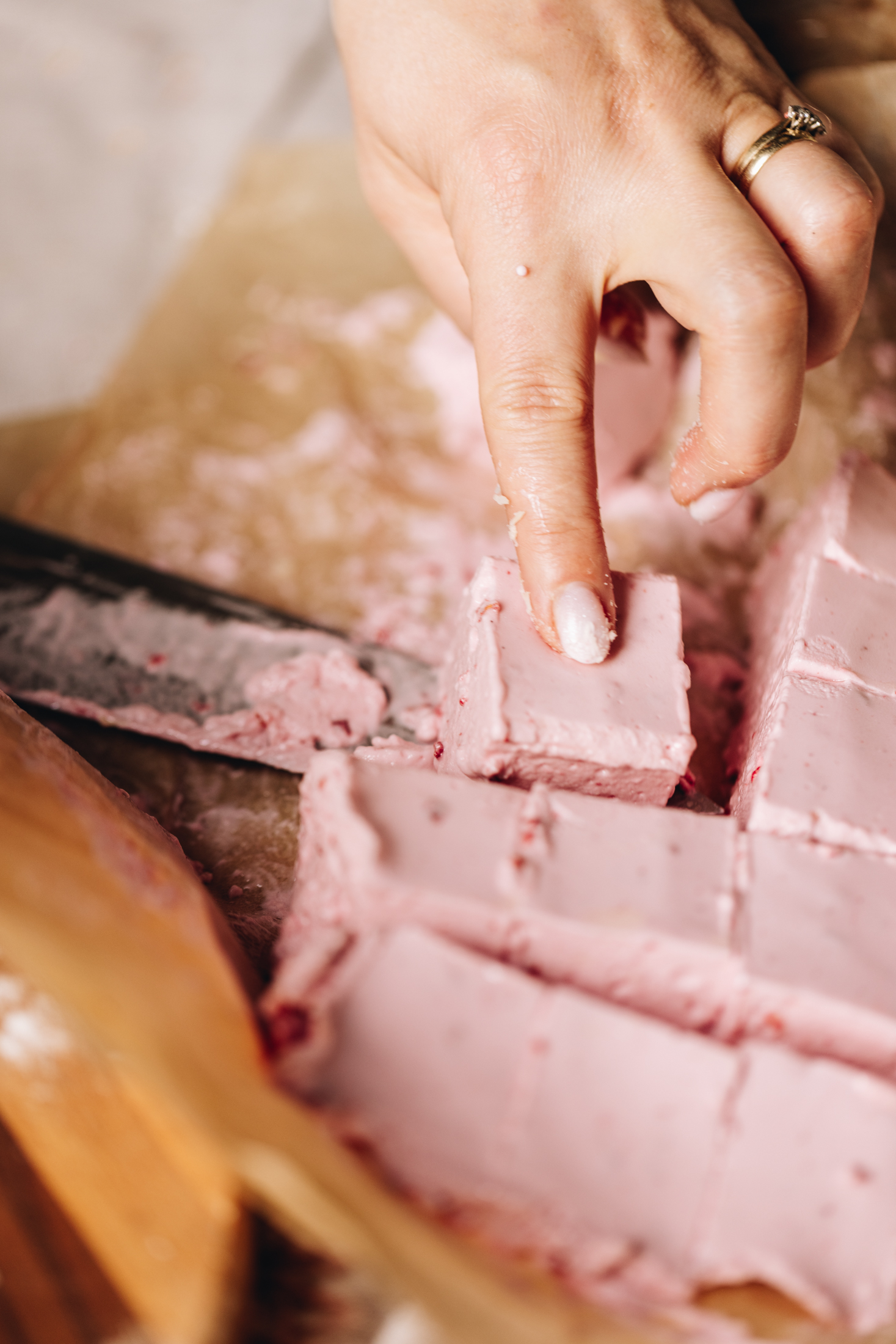 On brown baking paper is cut up slices of egg-free raspberry marshmallow. Naomi is using a large knife to remove one of the pieces. 