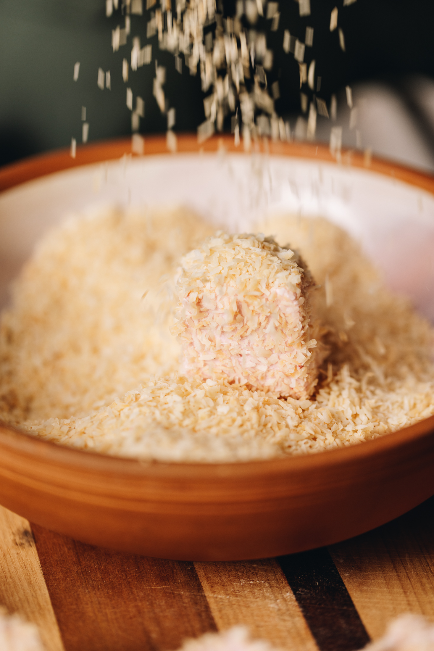 A brown ceramic bowl sits on a striped wooden board. In the bowl is toasted coconut and a square of marshmallow is  being covered with the coconut. Coconut is falling from the top of frame in to the bowl. 