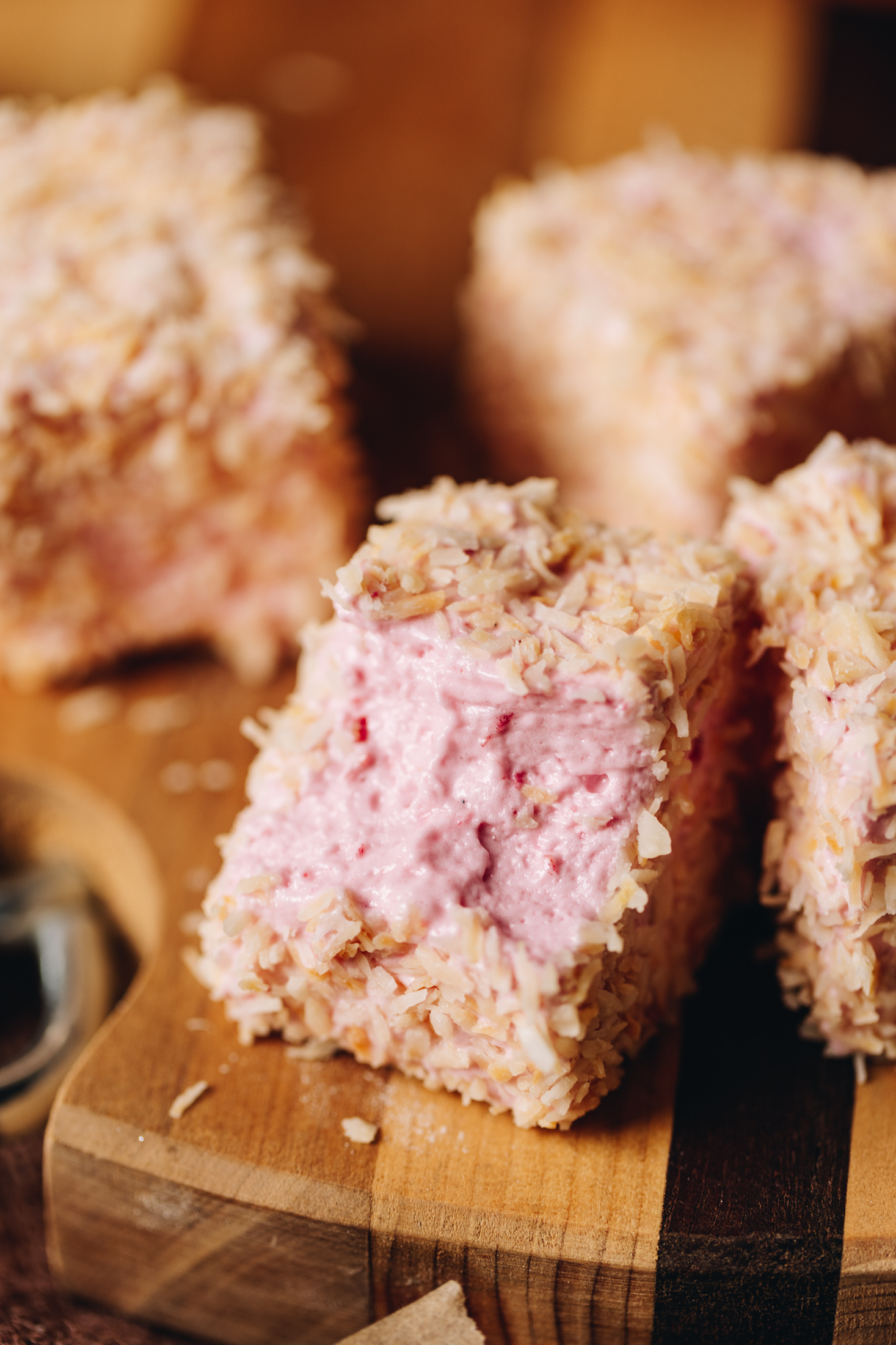 Squares of egg-free Raspberry marshmallow are on a striped wooden board. Each one is covered in toasted coconut. The front piece has bite mark in it.  