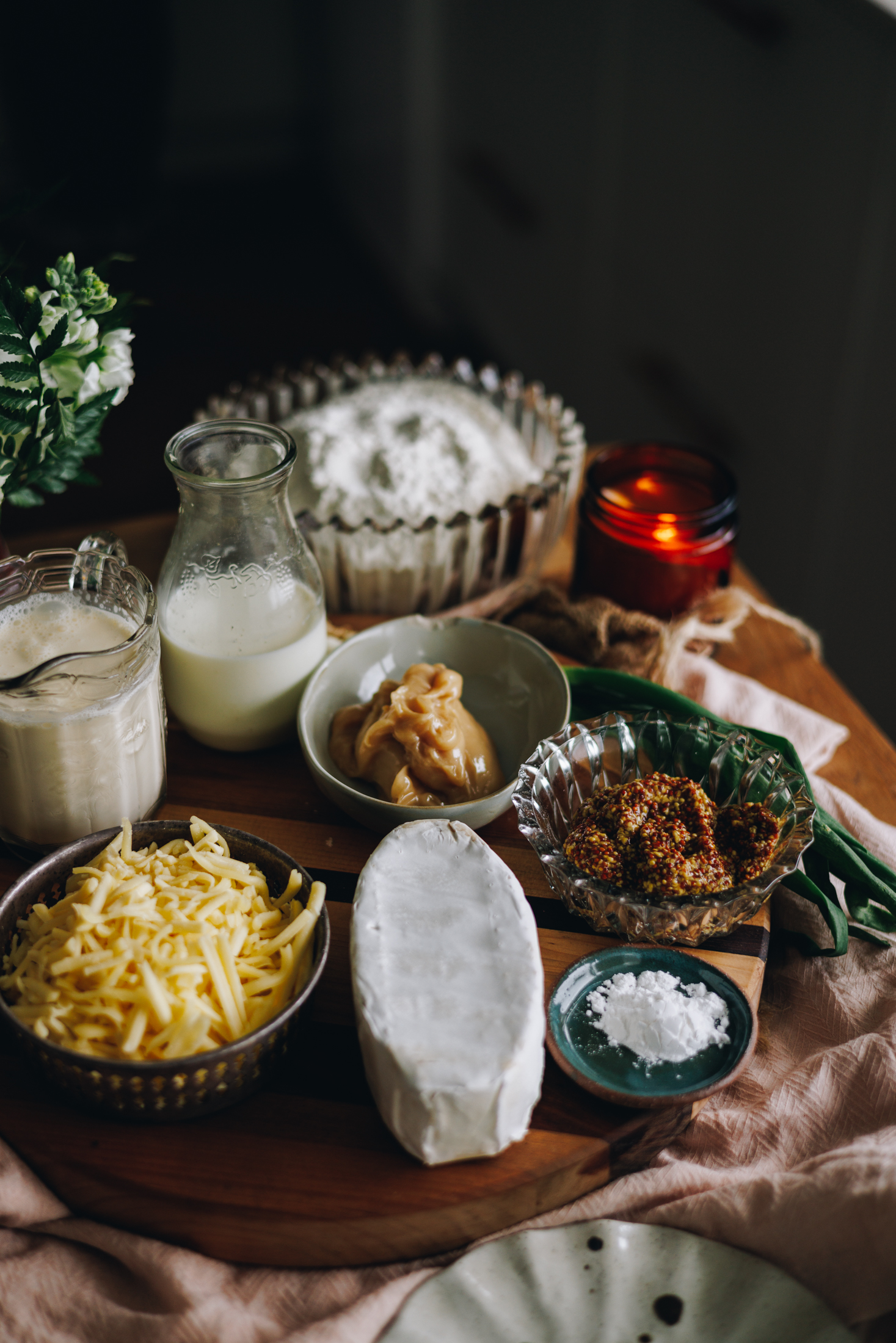 A wooden board sits on a pink tablecloth. On the board is vintage jars and bowls. There is brie cheese, mustard, honey, salt, grated cheese, milk, cream and flour in view. There is an amber candle burning in the background and green flowers in the corner.