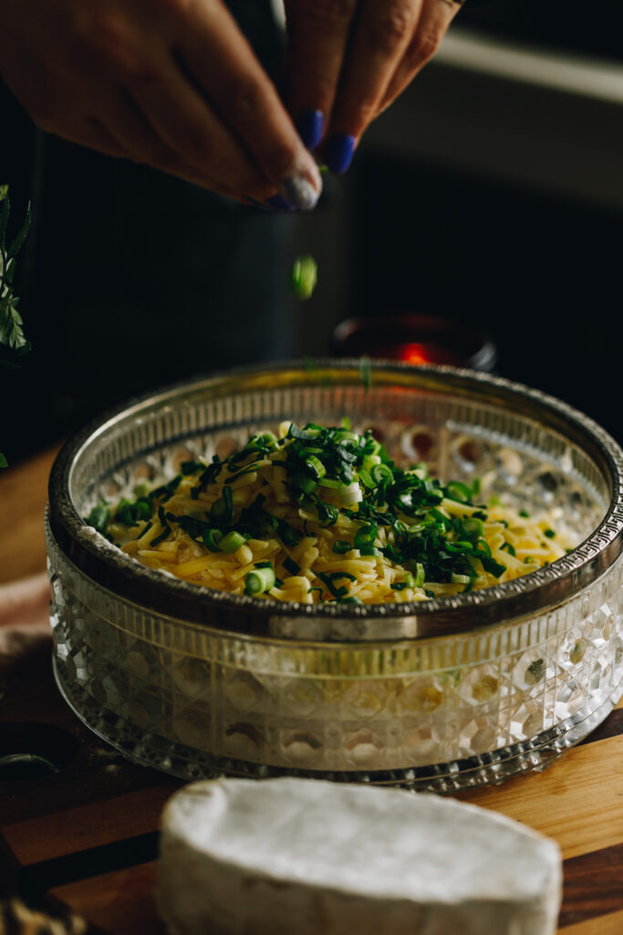 Sliced spring onions are being dropped in to a glass bowl with silver trim, the spring onions are falling in to grated cheese. A block of Brie Cheese is in front of it. The bowl is on a wooden board. 