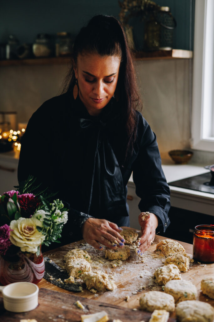 Naomi Toilalo is in her kitchen shaping the Brie Stuffed Honey Mustard Scone dough with her hands. Pieces of scone dough are all over the wooden table with a vase of flowers next to her. 