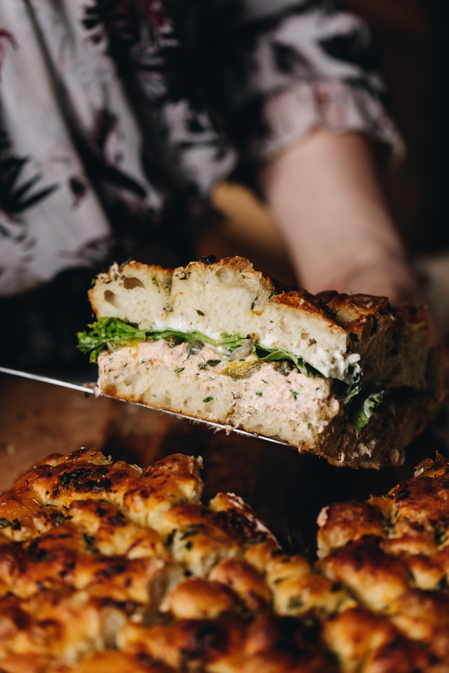 A large slice of creamy Salmond foccacia sandwich is held up. It shows the layers of fluffy focaccia brea, creamy salmon dip, lettuce and pickles.