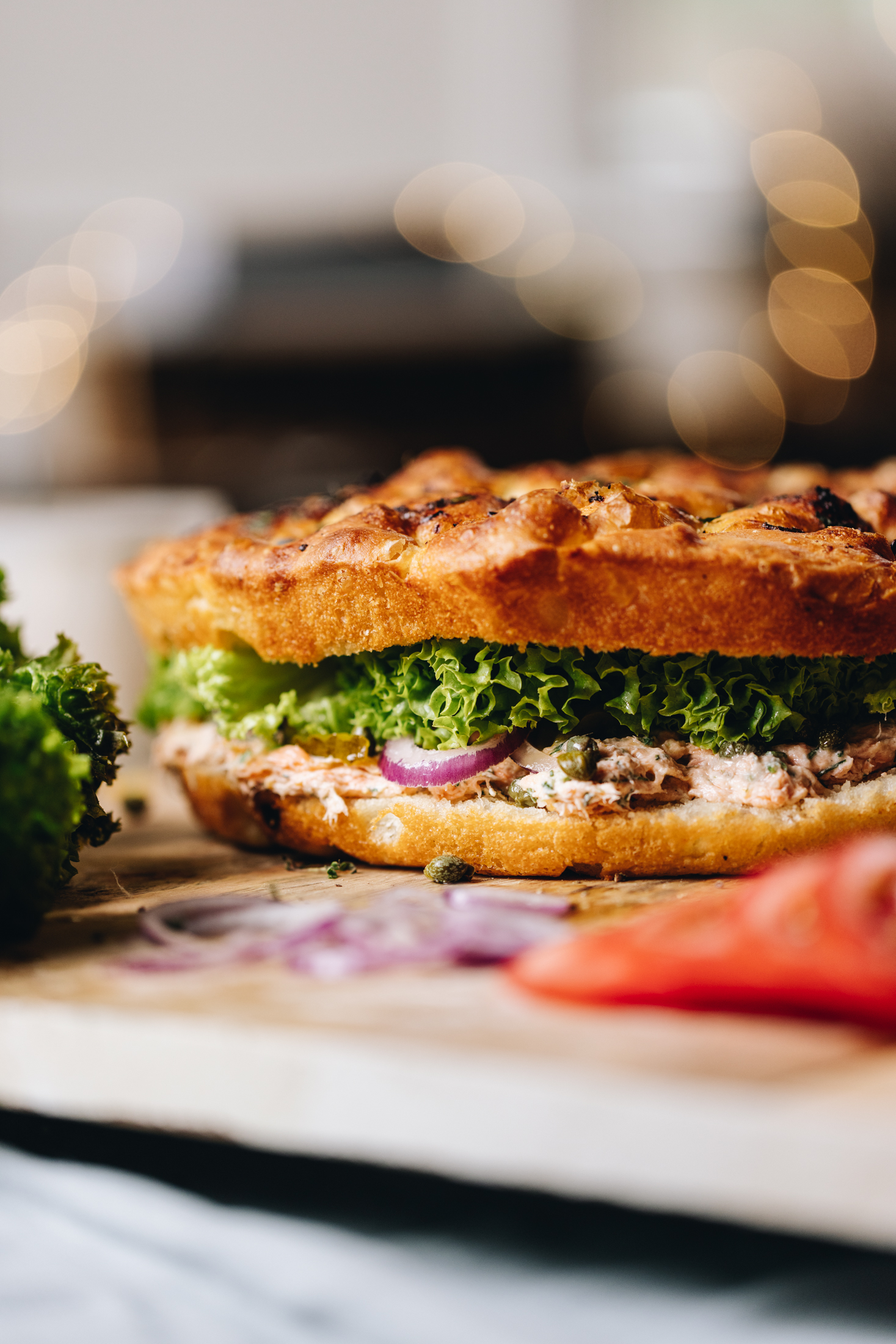 A whole creamy salmon focaccia sandwich sits on a wooden board. Salmon dip, capers, red onion and lettuce are seen in the sandwich. In front of it is more red onion, smoked Salmon and lettuce.