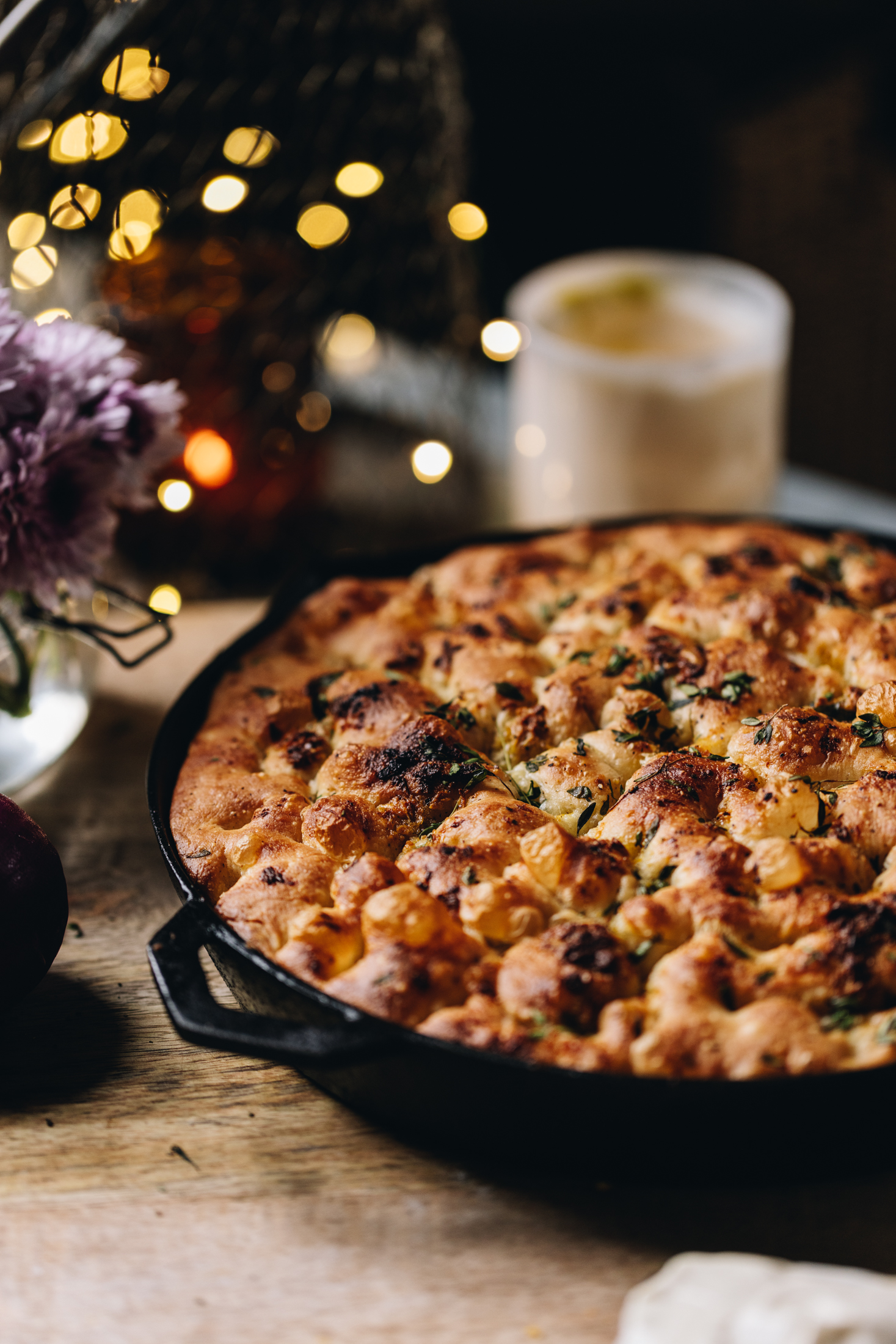 On a wooden table sits a freshly baked no-knead-focaccia bread that has been baked in a cast iron pan. Flowers, a candle and fairy lights are seen in the background.