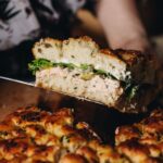 A piece of creamy salmon focaccia sandwich is held up in the air to show the insides of the sandwich, revealing salmon dip, pickles and lettuce.