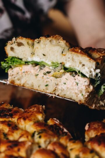 A piece of creamy salmon focaccia sandwich is held up in the air to show the insides of the sandwich, revealing salmon dip, pickles and lettuce.