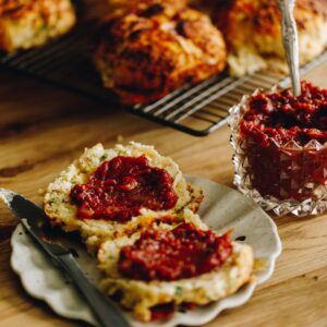 easy tomato chutney is spread on to baked scones that are split open on a plate. More chutney is seen in the glass jar behind the plate.