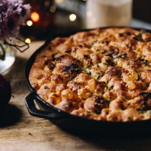 No-knead focaccia bread is baked in a cast iron pan with bubbles all over top. It is sitting on a wooden board.