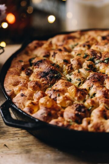 No-knead focaccia bread is baked in a cast iron pan with bubbles all over top. It is sitting on a wooden board.