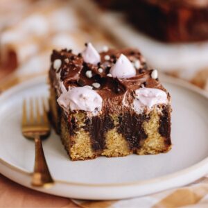 A square slice of chocolate ganache poke cake is on a white plate with a gold fork next to it. The ganache in the cake is seen and it is decorate with pink meringue blobs and sprinkles.