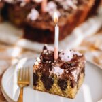 A square slice of chocolate ganache poke cake is on a white plate with a gold fork next to it. The ganache in the cake is seen and it is decorate with pink meringue blobs and sprinkles. It has a single candle in it that is lit.