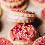 A close up shows rustic, homemade 100's and 1000's cookies with pink icing and sprinkles. One of them sits on an angle, revealing the centre that has been filled with pink marshmallow.