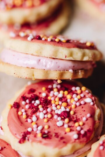 A close up shows rustic, homemade 100's and 1000's cookies with pink icing and sprinkles. One of them sits on an angle, revealing the centre that has been filled with pink marshmallow.