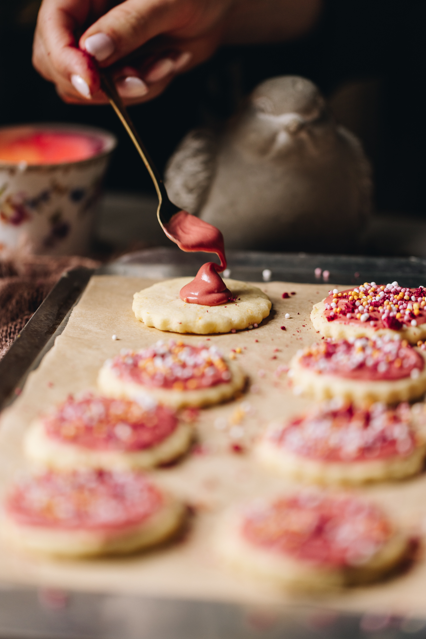 On a black tray with brown baking paper is hundreds and thousands biscuits baked, a spoon is topping pink white chocolate on top. The rest of the tray if filled with decorated biscuits.