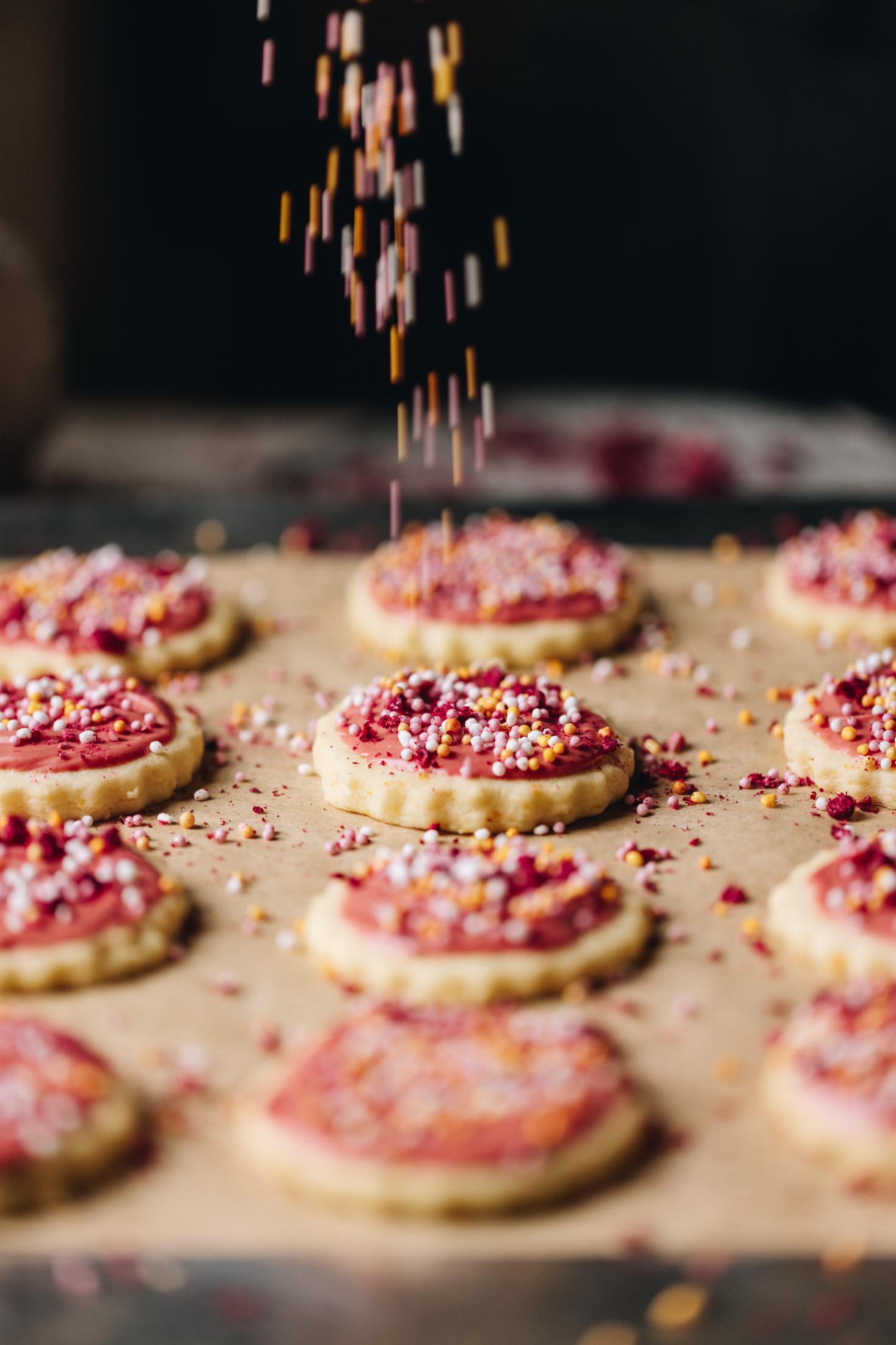 On a black tray with brown baking paper is hundreds and thousands biscuits baked, sprinkles are falling from above on to one of the biscuits.