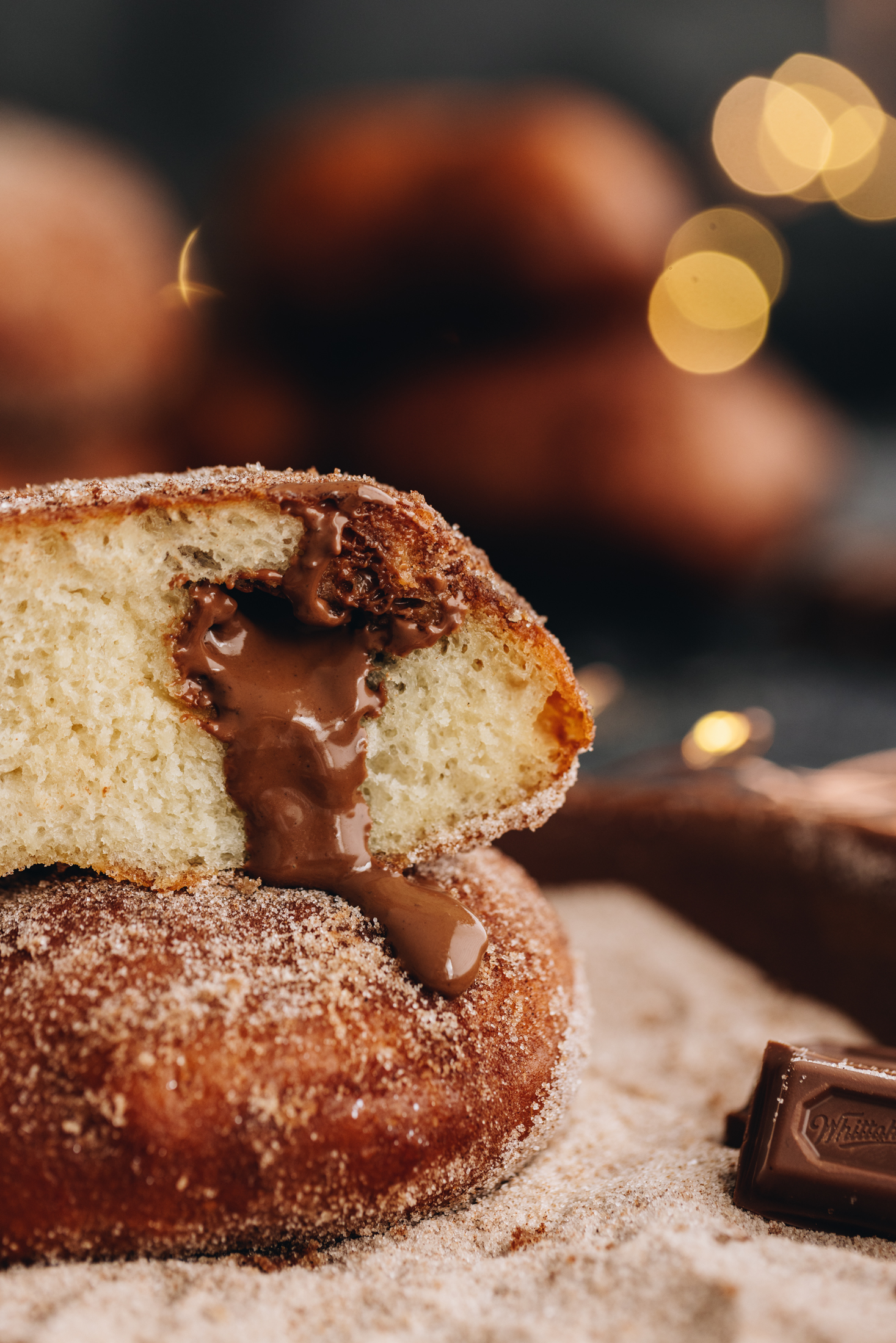 Two pieces of chocolate stuffed fry bread are stacked on top of each other. One of the faces the back and the other reveals the gooey chocolate centre coming out of the dough. The fry bread is covered in cinnamon sugar and is sitting in a bowl of cinnamon sugar with a piece of chocolate next to it. Fairy lights glow in the background. 