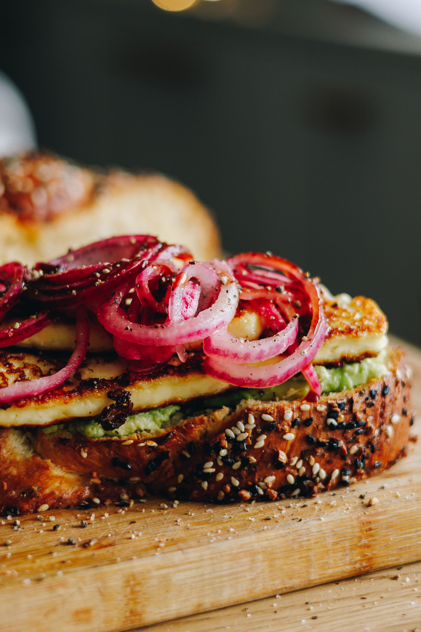 A wooden board sits on a wooden table. On the board is a freshly baked Savoury braided brioche bread loaf that has been cut open. In front of it is a freshly made avocado and halloumi sandwich. A slice of the bread has avocado smeared on to it with grilled halloumi pieces and is piled with red pickled onions and loads of cracker pepper. 