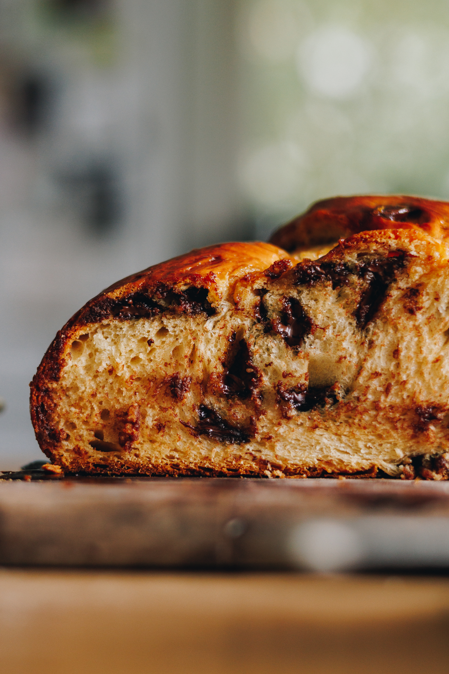 On a wooden table sits a wooden chopping board. On the board is a freshly baked Braided Chocolate Chip bread that has been cut open to reveal the gooey chocolate and soft bread. 