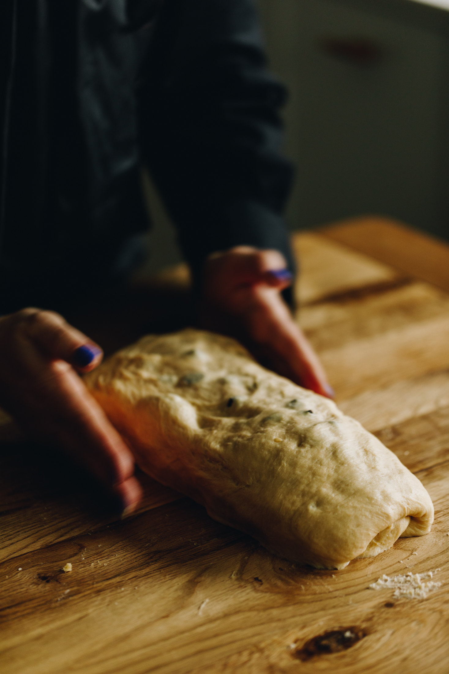 On a wooden table is a braided chocolate chip bread dough that is being kneading with Naomi's hands. In the dough speckles of chocolate chop drops are seen. 