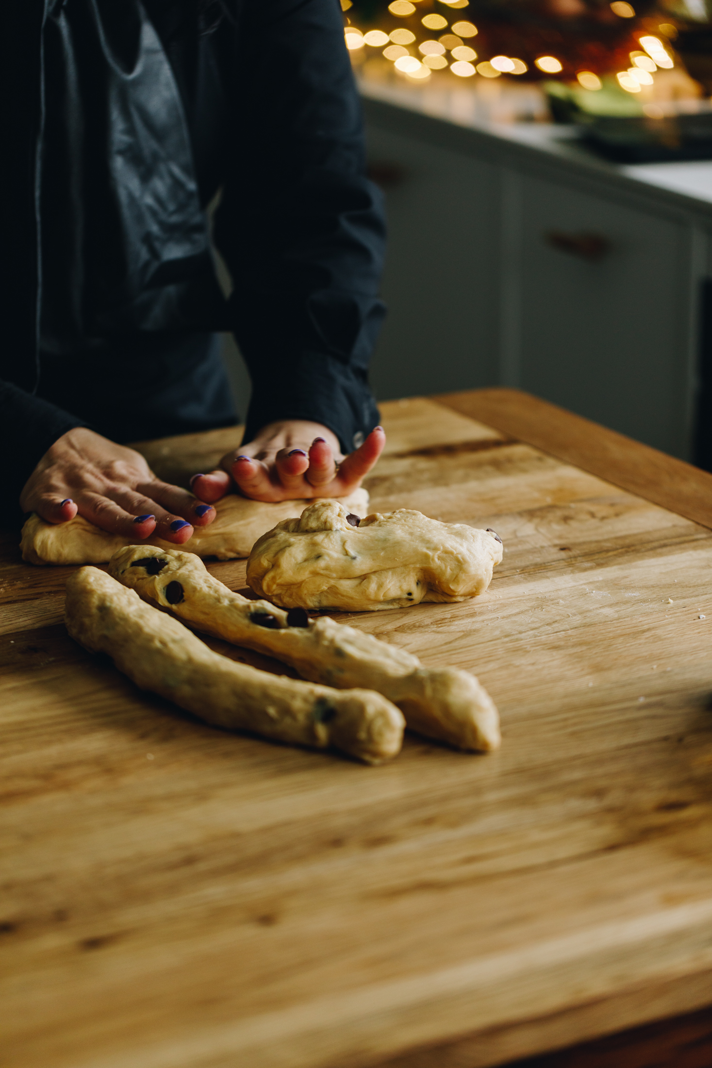 On a wooden table is a braided chocolate chip bread dough. It has been separated in to four pieces. Two pieces have been rolled in to logs, one is still a blob and the other is being rolled in to a long with Naomi's hands. In the dough speckles of chocolate chop drops are seen.