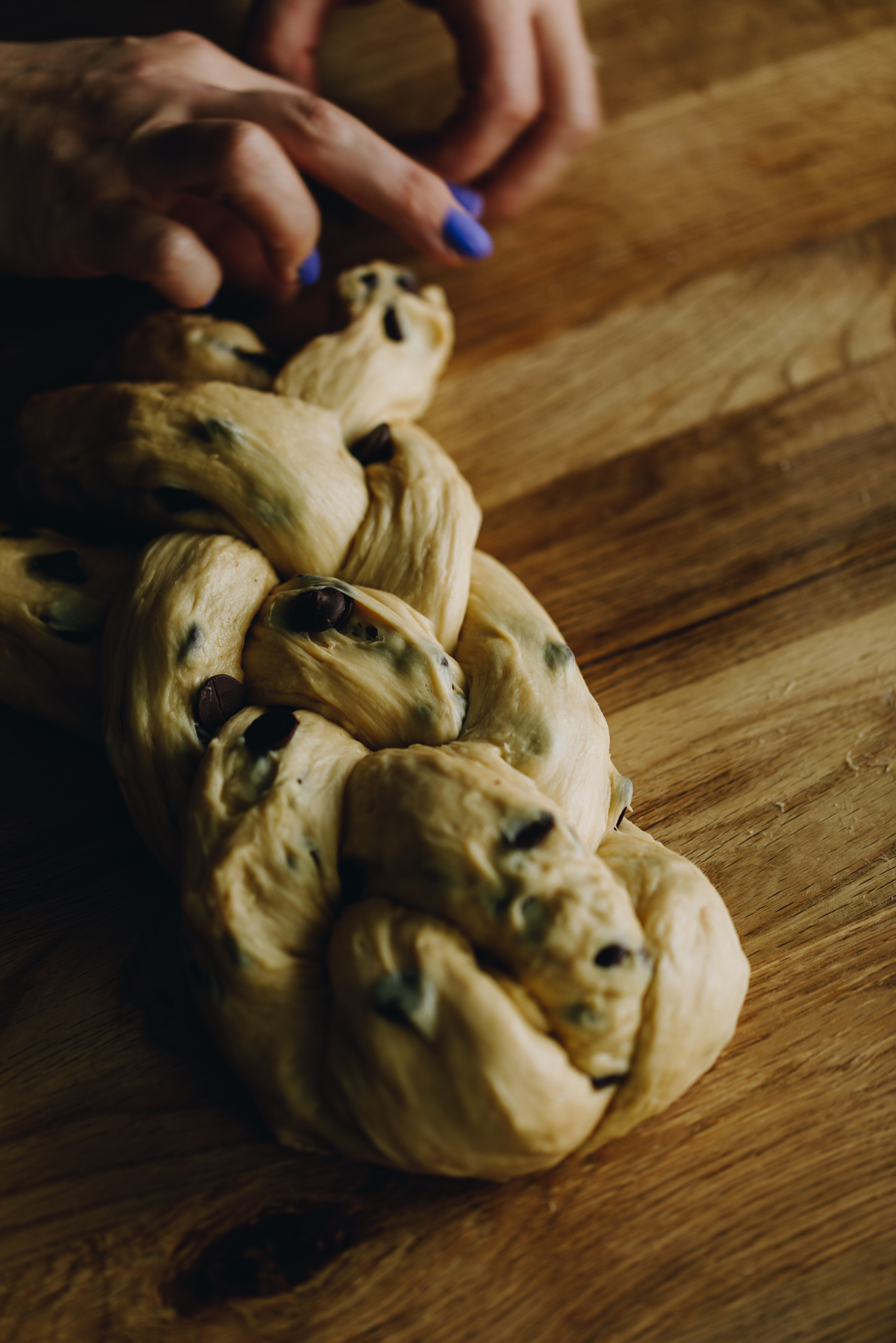 On a wooden table is a braided chocolate chip bread dough. It is being braided with four strands of dough with Naomi;'s hands. In the dough speckles of chocolate chop drops are seen.