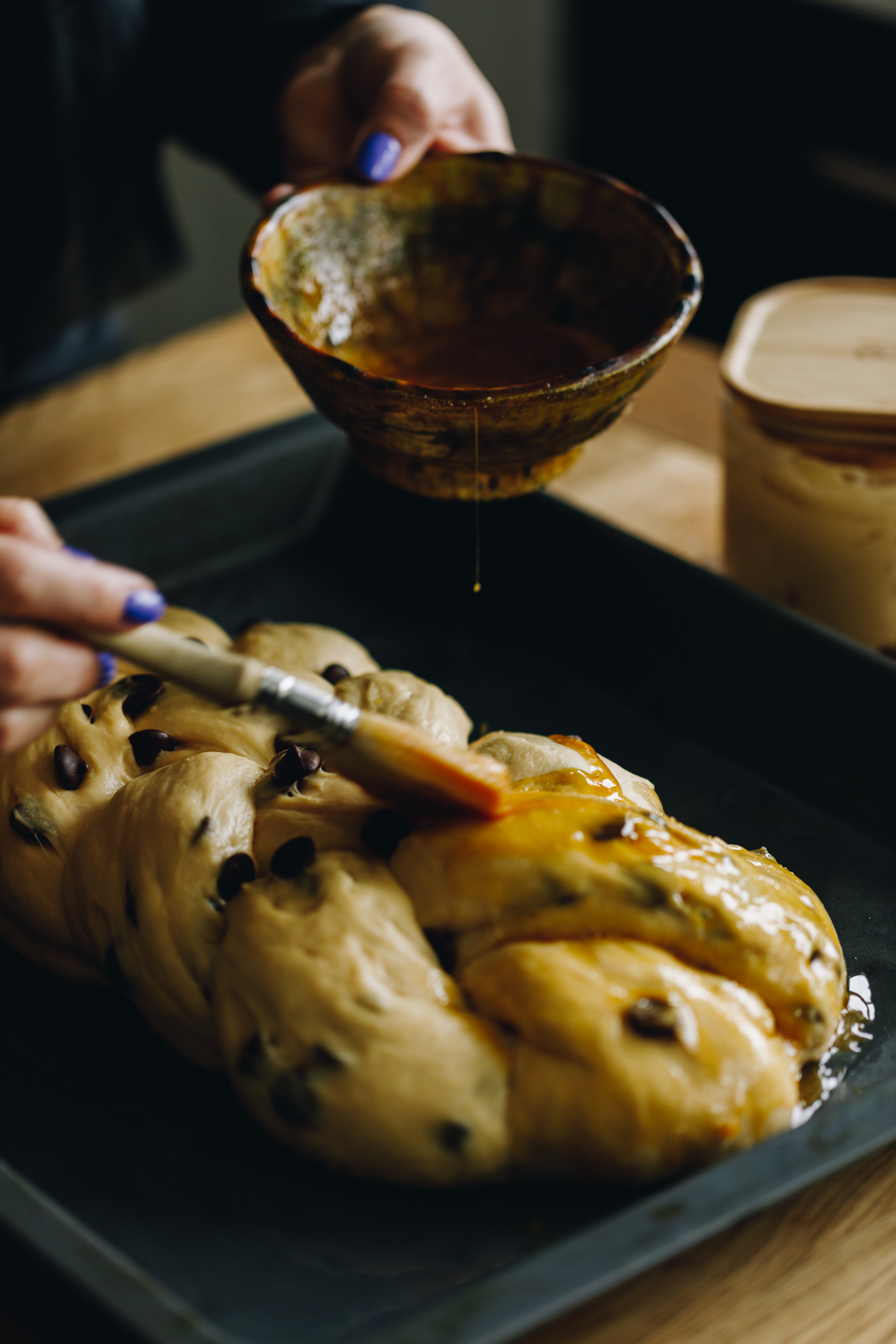 Braided chocolate chip dough has been braided in to a four plait and sits on a black tray that is on a wooden table. In a green ceramic bowl is an egg mixture, Naomi is brushing the mixture to the bread dough with a pastry brush. 