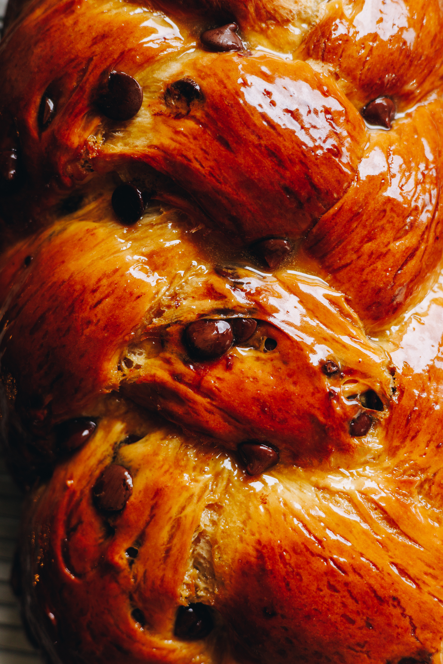 A close up flat lay shot shows a freshly baked braided chocolate chip bread. It is deeply golden and has been covered in a maple syrup and butter glaze. The bread has been braided and chocolate drops are studded all over it in the dough. 