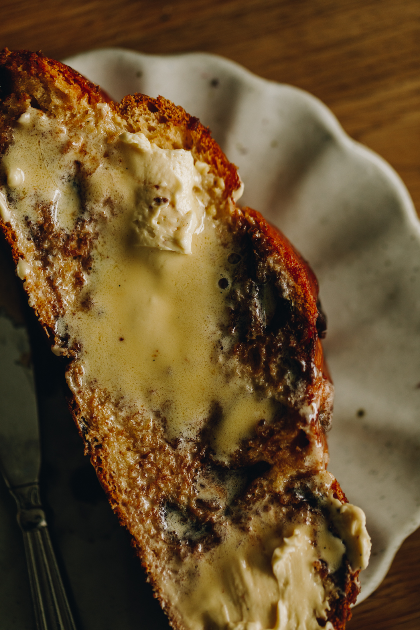 A piece of freshly baked braided chocolate chip bread is on a ceramic frilled plate that is sitting on a wooden table. It has been smeared with whipped butter and it is melting in to the bread. 