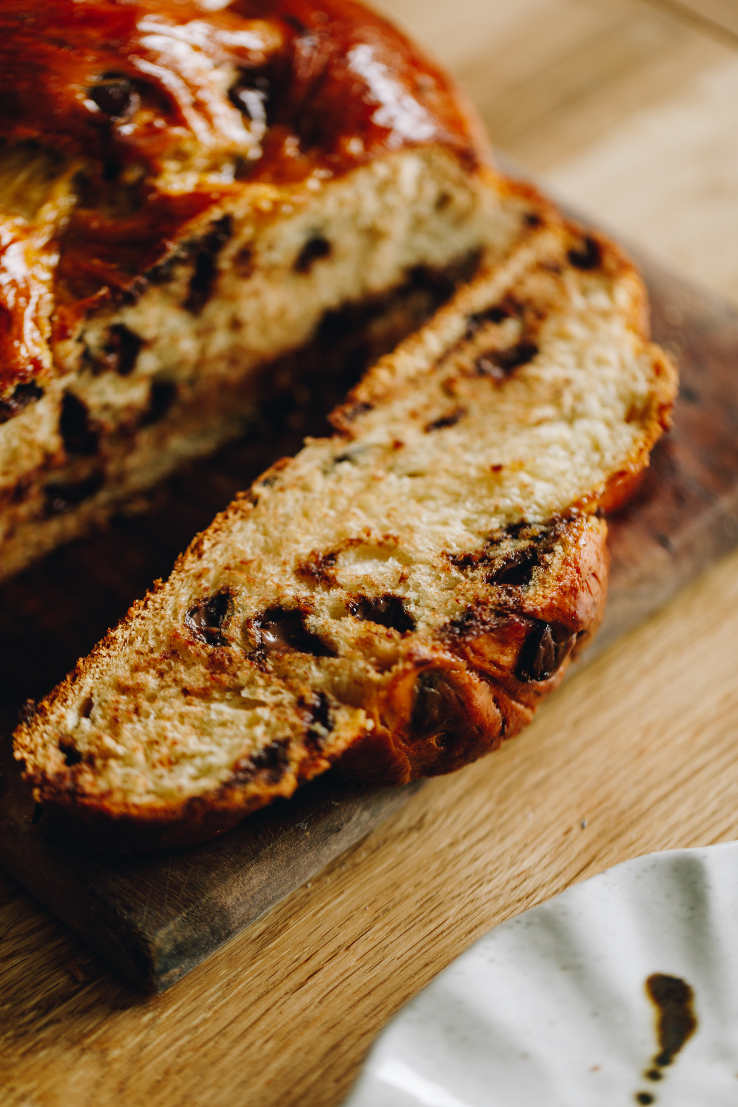 On a wooden table sits a wooden chopping board. On the board is a freshly baked Braided Chocolate Chip bread that has been cut open to reveal the gooey chocolate and soft bread. One slice lays down on the wooden board. The remaining loaf is glossy. 