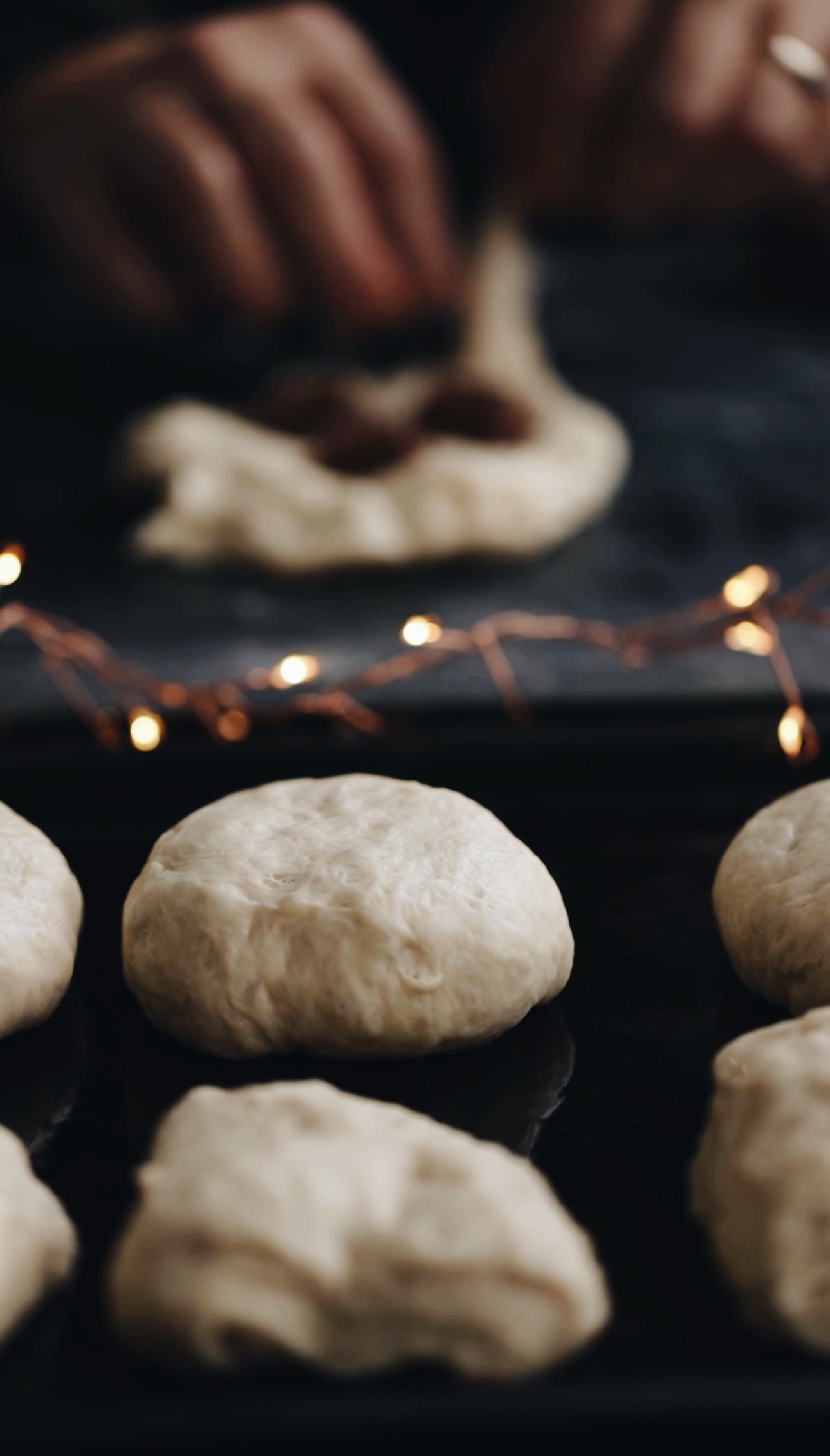 A black tray has fry bread on it with golden fairy lights behind it. Behind the fry bread Naomi is stuffing a piece of dough with chocolate. 