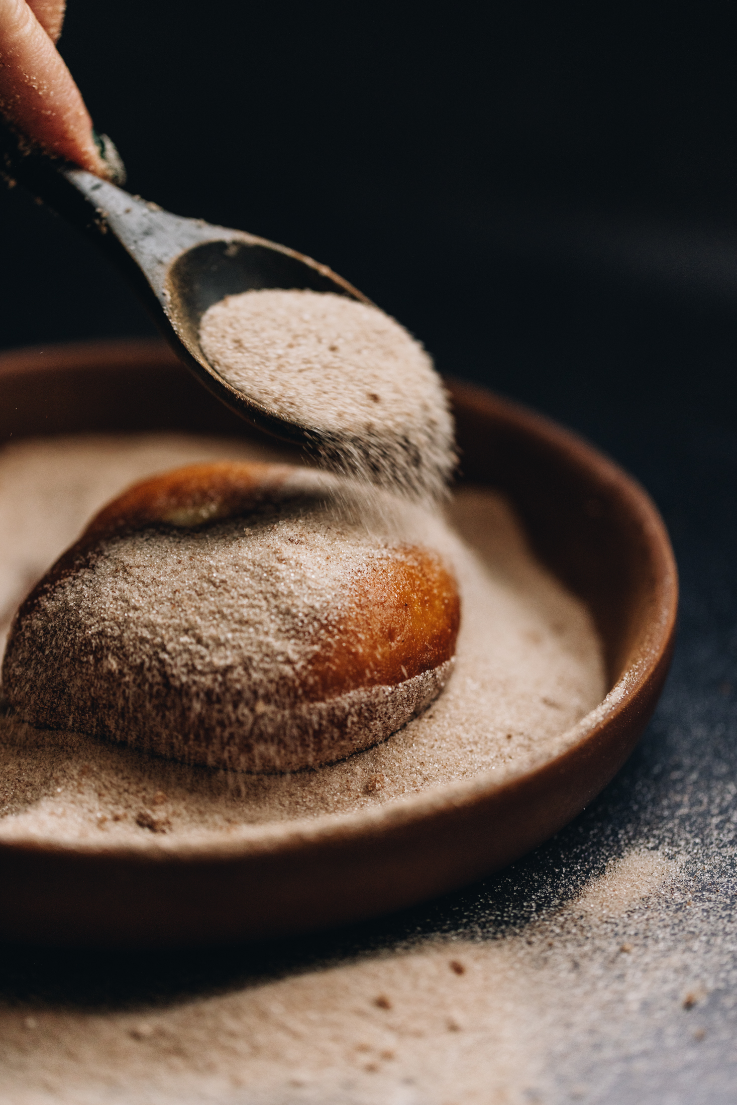 A round wooden bowl sits on a black bench. In the bowl is cinnamon sugar and a wooden spoon is adding cinnamon sugar on top of a chocolate stuffed fry bread. There is cinnamon sugar on the bench too. 