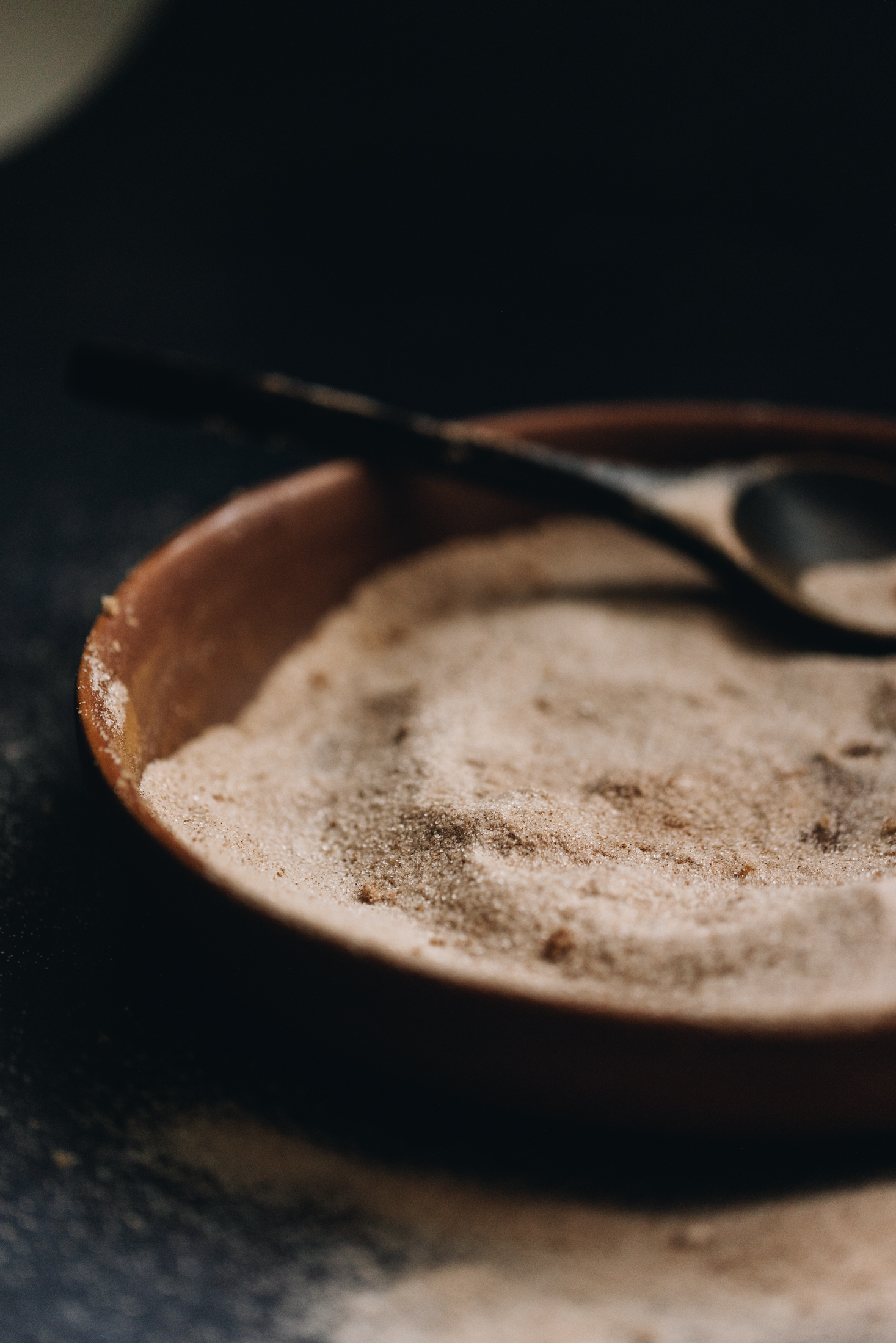 A brown wooden bowl sits on a dark bench. In the bowl is a spiced sugar with some of it sprinkled on the bench. In the bowl is a black wooden spoon.