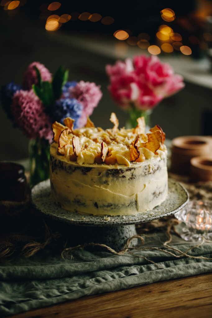 A ceramic cake stand sits on a wooden table with two bunches of flowers in the background. On the cake stand is a Sticky Caramel Date Cake with cream cheese frosting and apple chips on top. 