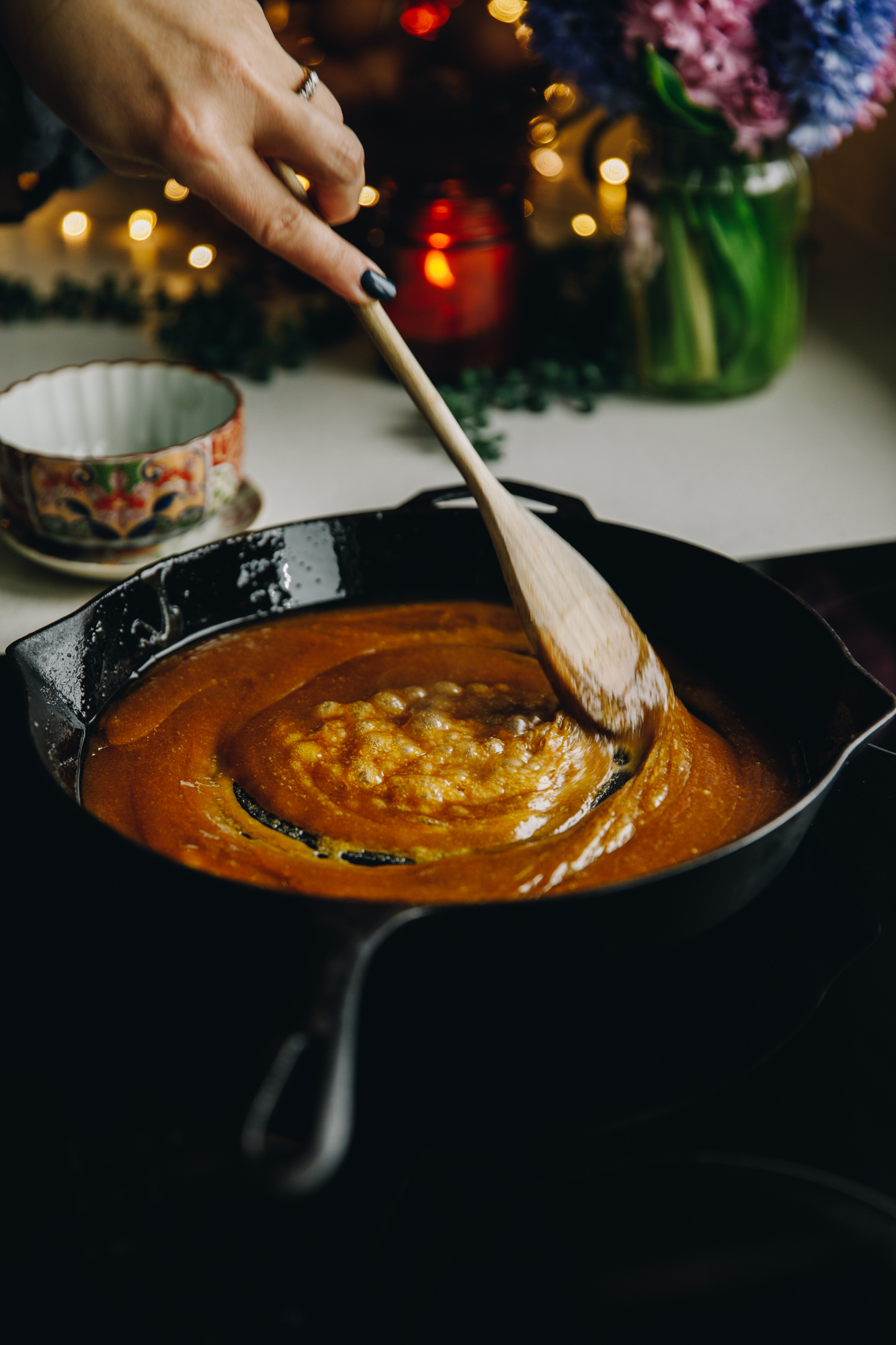 On a stove sits a cast iron pan with bubbly camrale in it. A wooden spoon is stirring it as it bubbles. There is flowers and a candle on the bench behind it.