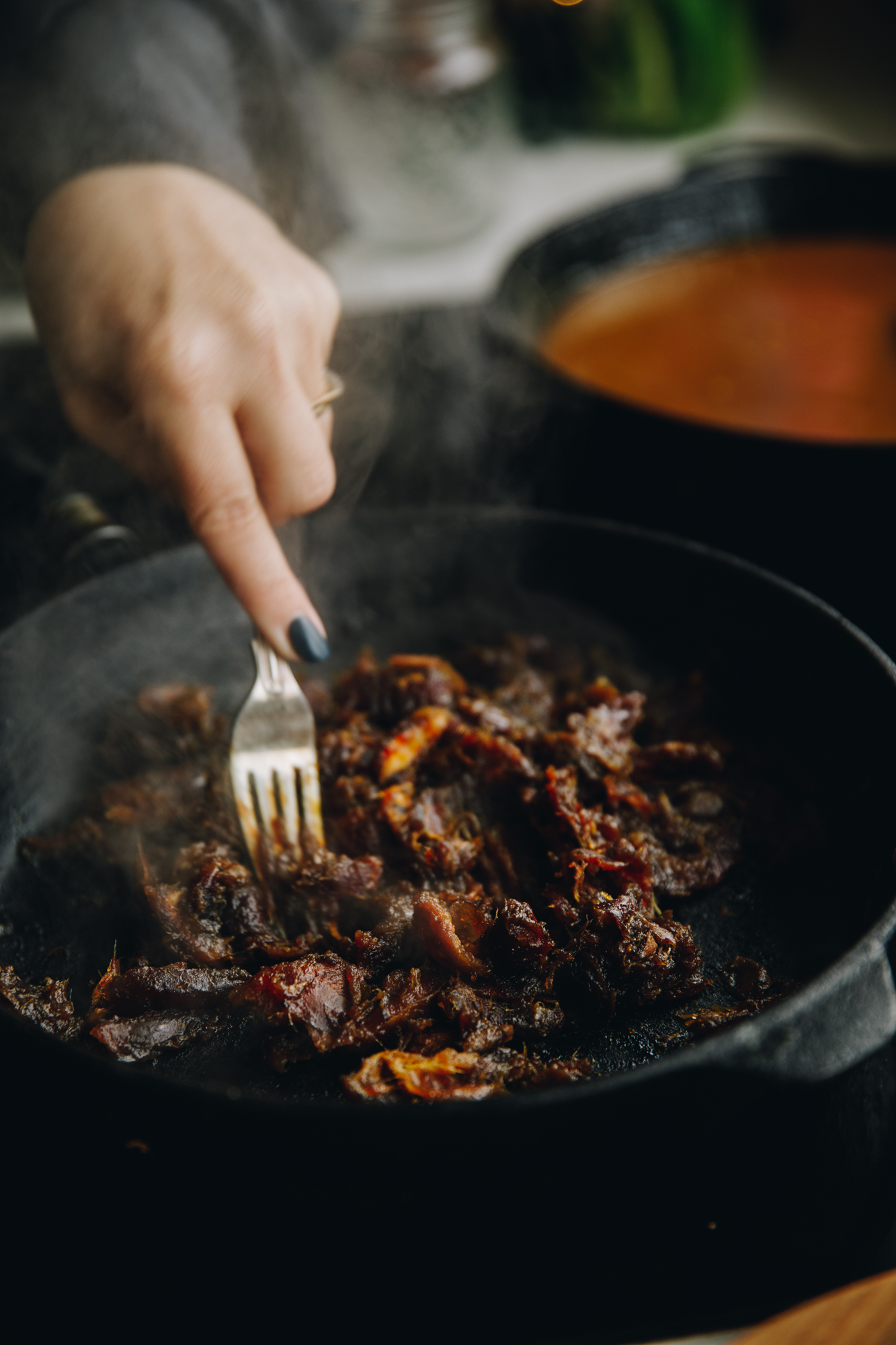 On a stove sits a cast iron pan with dates on it, they are steaming hot and are being mashed with a fork. A pan of caramel is on the stove behind it. 