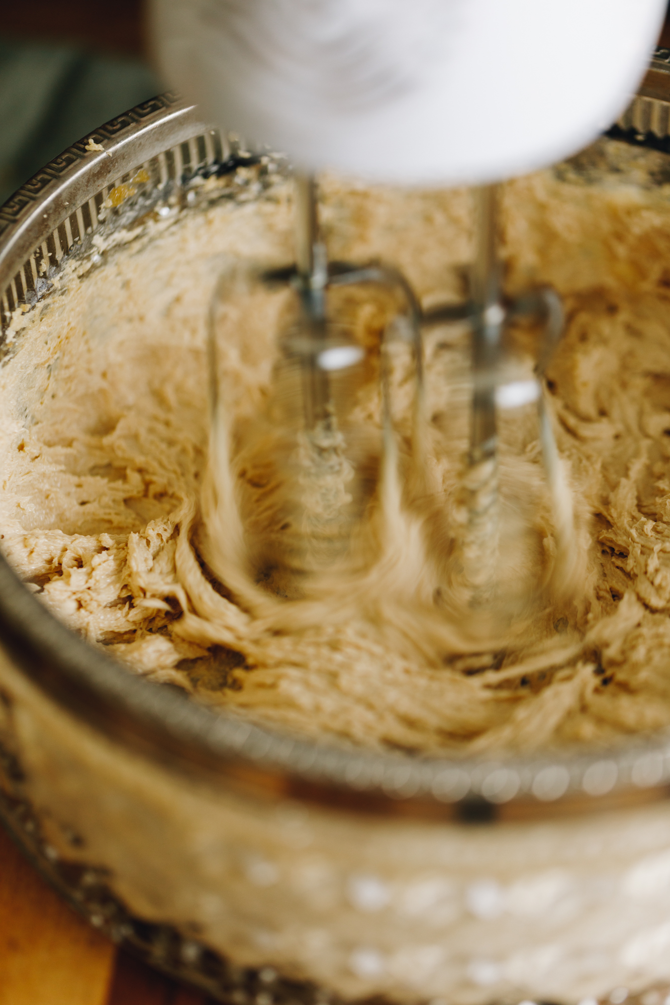 In a vintage glass bowl is butter and sugar being whipped with a w white hand mixer. It is sitting on a wooden board. 