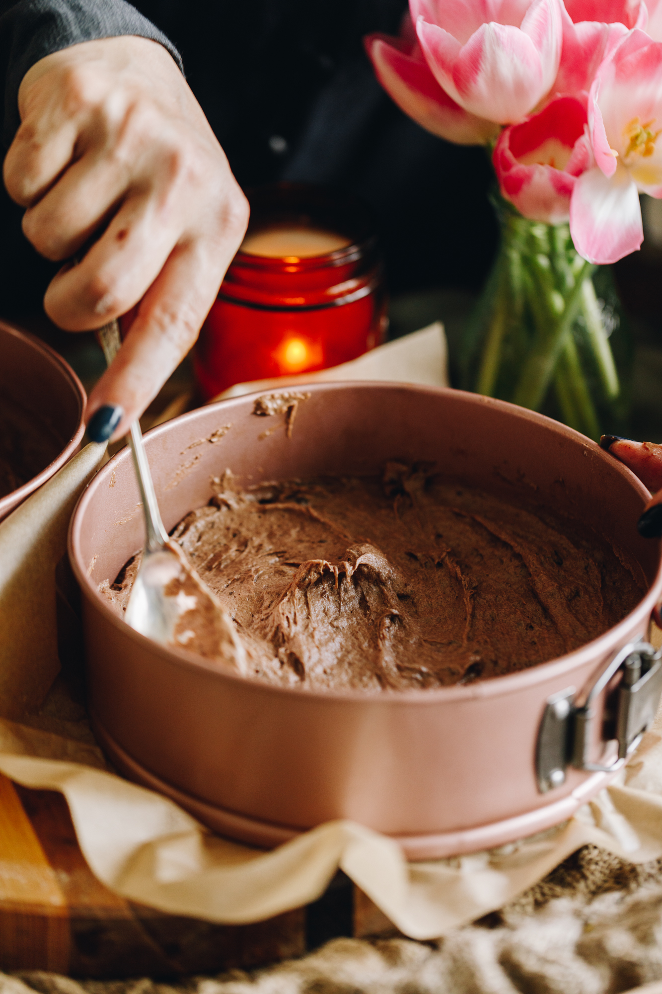 A rose pink cake tin sits on a wooden table with a candle in the background. In the cake tin is unbaked cake mixture that is being spread with a spoon. Pink flowers are in the background. 
