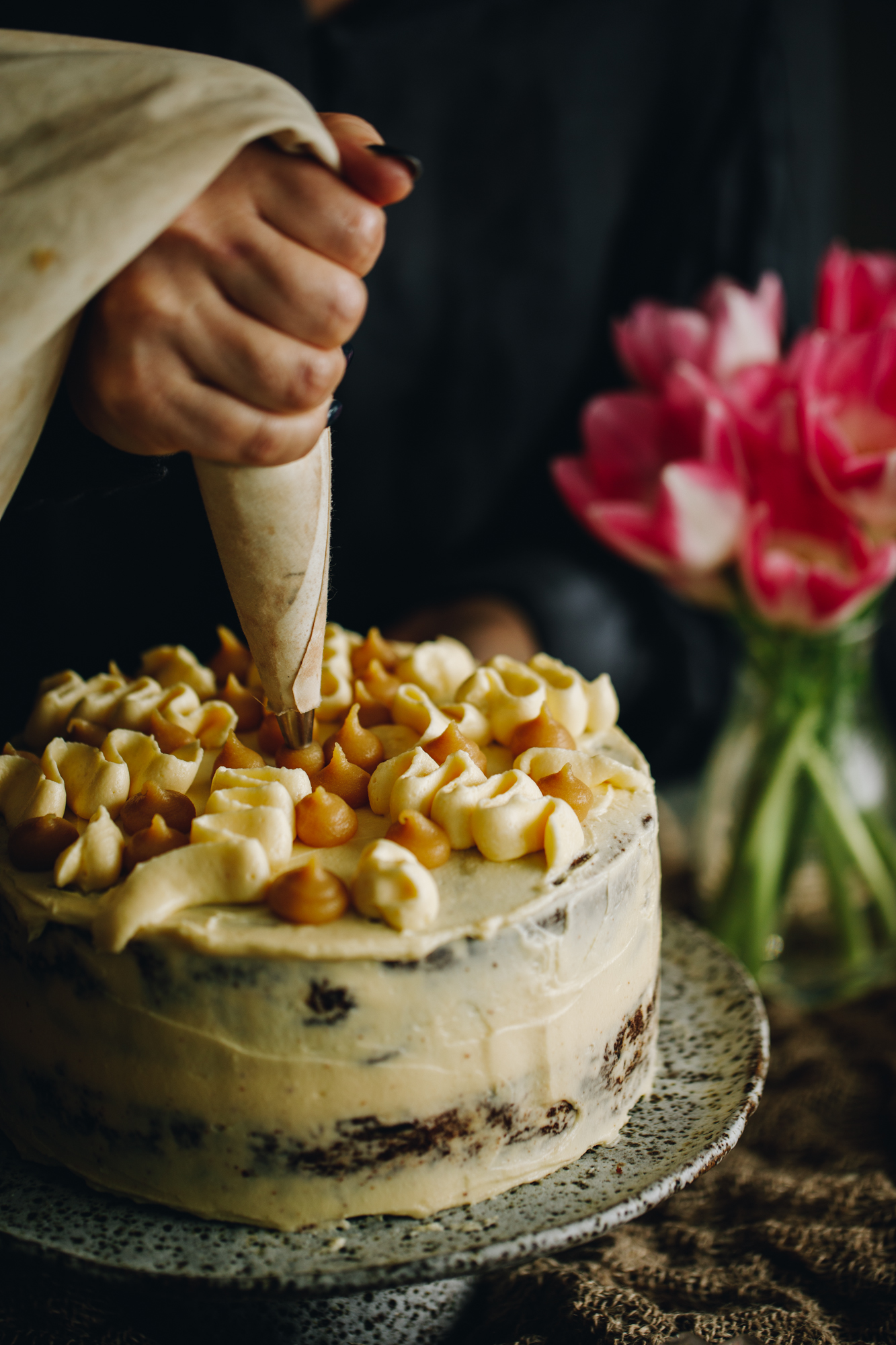 A ceramic cake stand sits on a wooden table with a bunch of flowers in the background. On the cake stand is a Sticky Caramel Date Cake with cream cheese frosting piped on to it. Naomi is now piping caramel on to the cake with a piping bag.