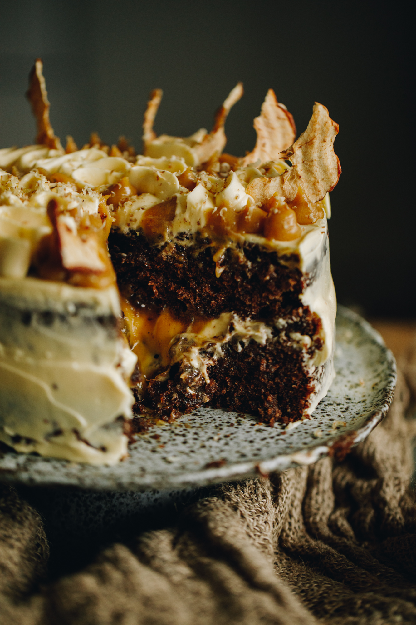 A ceramic cake stand sits on a wooden table On the cake stand is a Sticky Caramel Date Cake with cream cheese frosting and caramel piped on to it. It also has piece of dried apple crisps. The cake has been cut open and it reveals the gooey caramel and frosting oozing out of the centre. 