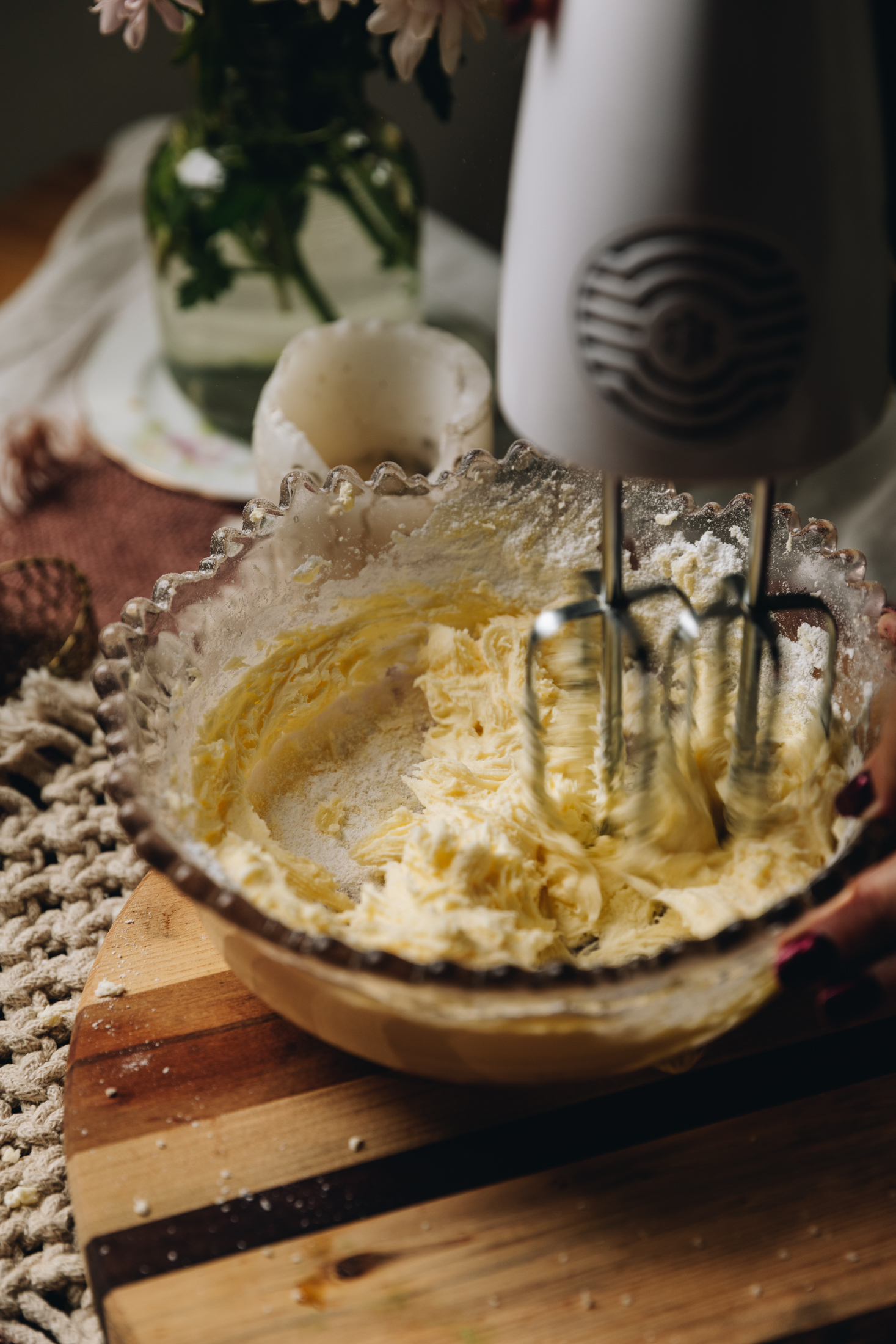In a vintage glass bowl is butter and sugar being whipped with a w white hand mixer. It is sitting on a wooden board.
