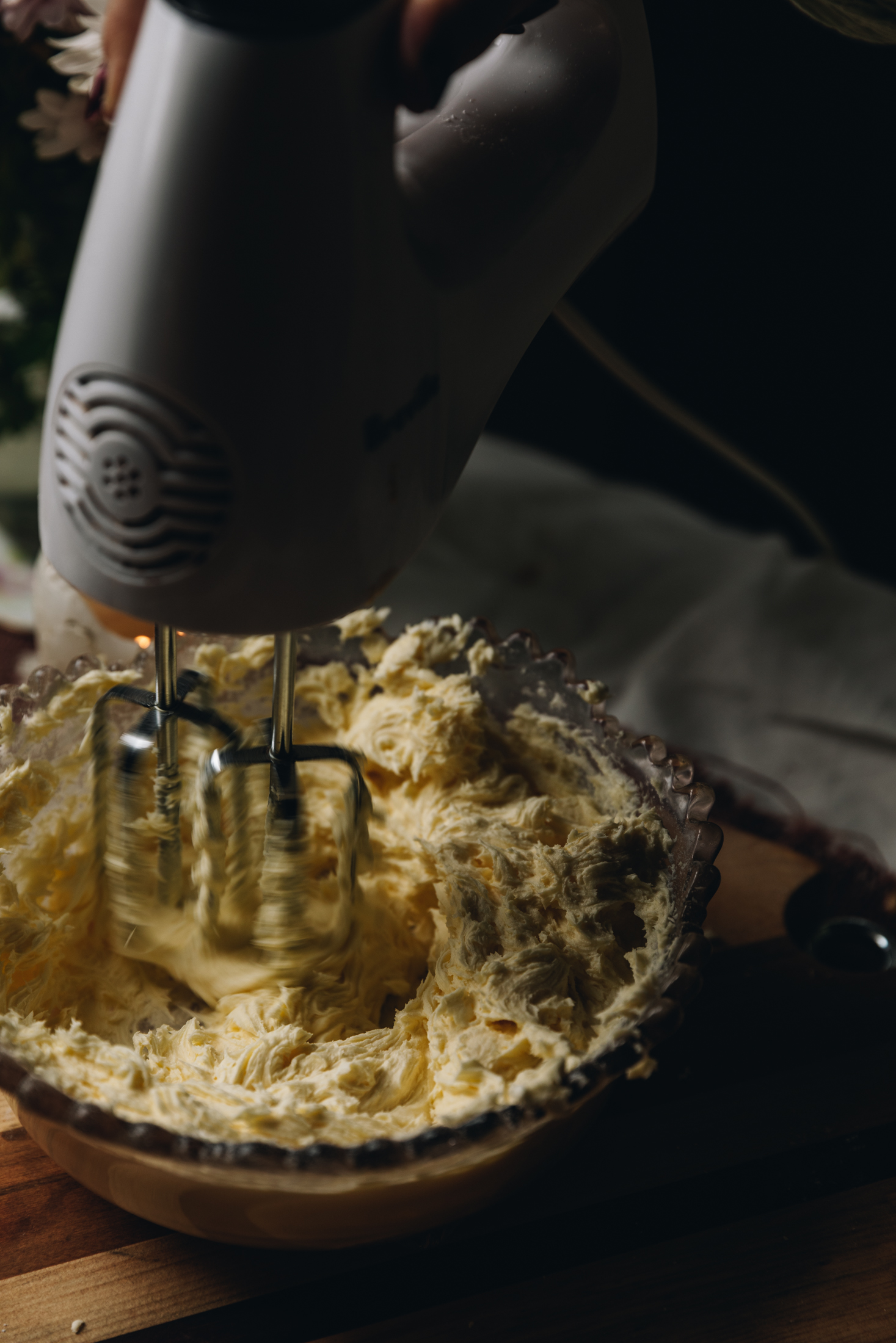 In a vintage glass bowl is cream cheese frosting being whipped with a white hand mixer. It is sitting on a wooden board.