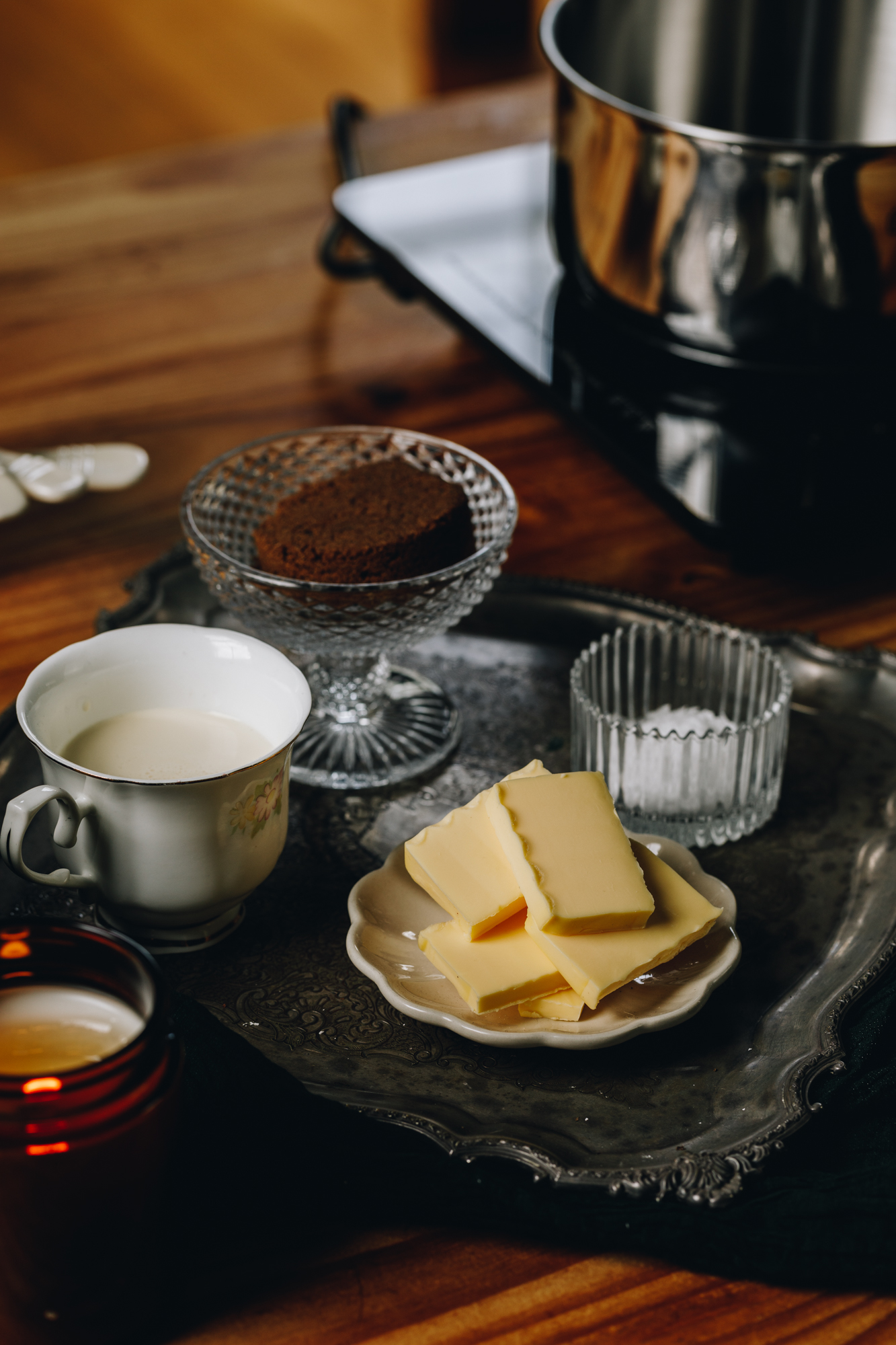 A silver vintage that is sitting on a wooden table. It has cream, brown sugar, butter and vanilla in vintage glasses and plate. There is a pot and a black portable stove in the background. 